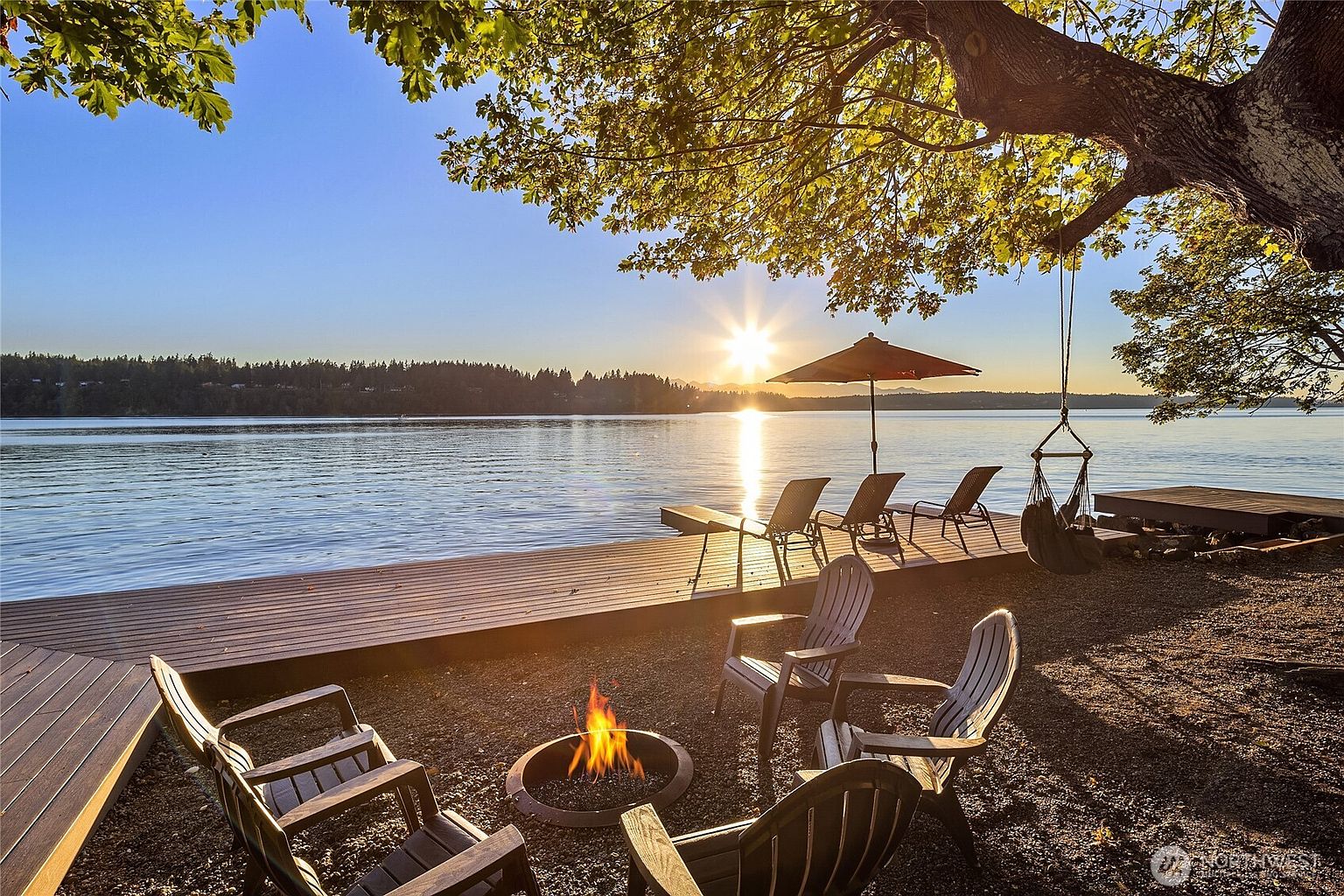 This image showcases a serene waterfront patio and deck area, perfect for relaxation and entertainment. A fire pit surrounded by chairs creates a cozy gathering space, while lounge chairs on the deck offer stunning water views. The scene is bathed in warm sunlight, enhancing the inviting atmosphere and highlighting the property's outdoor living potential.