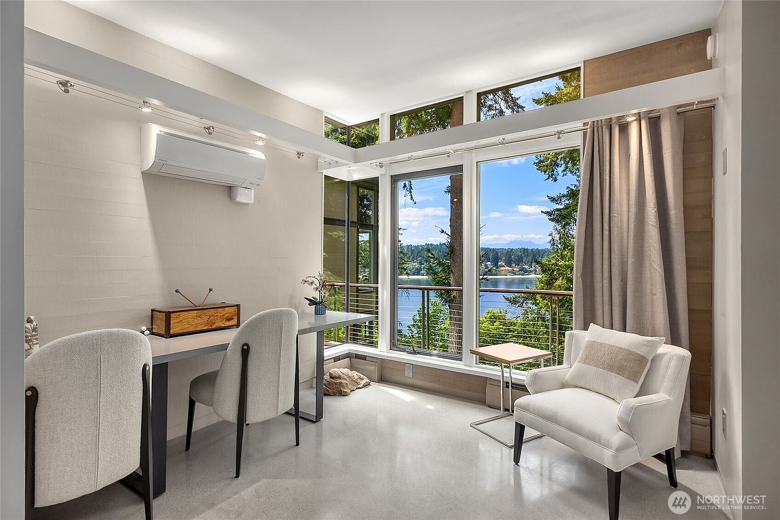 This is a bright and modern office space with a stunning view of the water. The room features a sleek desk with two chairs, a comfortable armchair, and large windows that let in plenty of natural light. The neutral color palette and minimalist design create a calming and productive atmosphere.