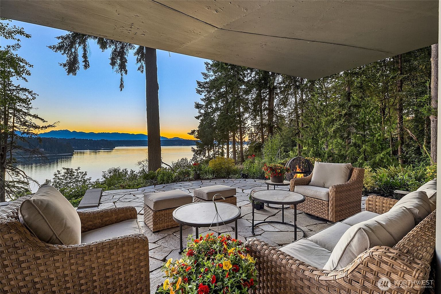 This image showcases a luxurious outdoor patio area with comfortable wicker furniture, including armchairs, ottomans, and a sofa, arranged around coffee tables. The patio is paved with natural stone and offers stunning views of a serene lake and distant mountains at sunset. A fire pit adds warmth and ambiance to the space, surrounded by lush greenery and trees.