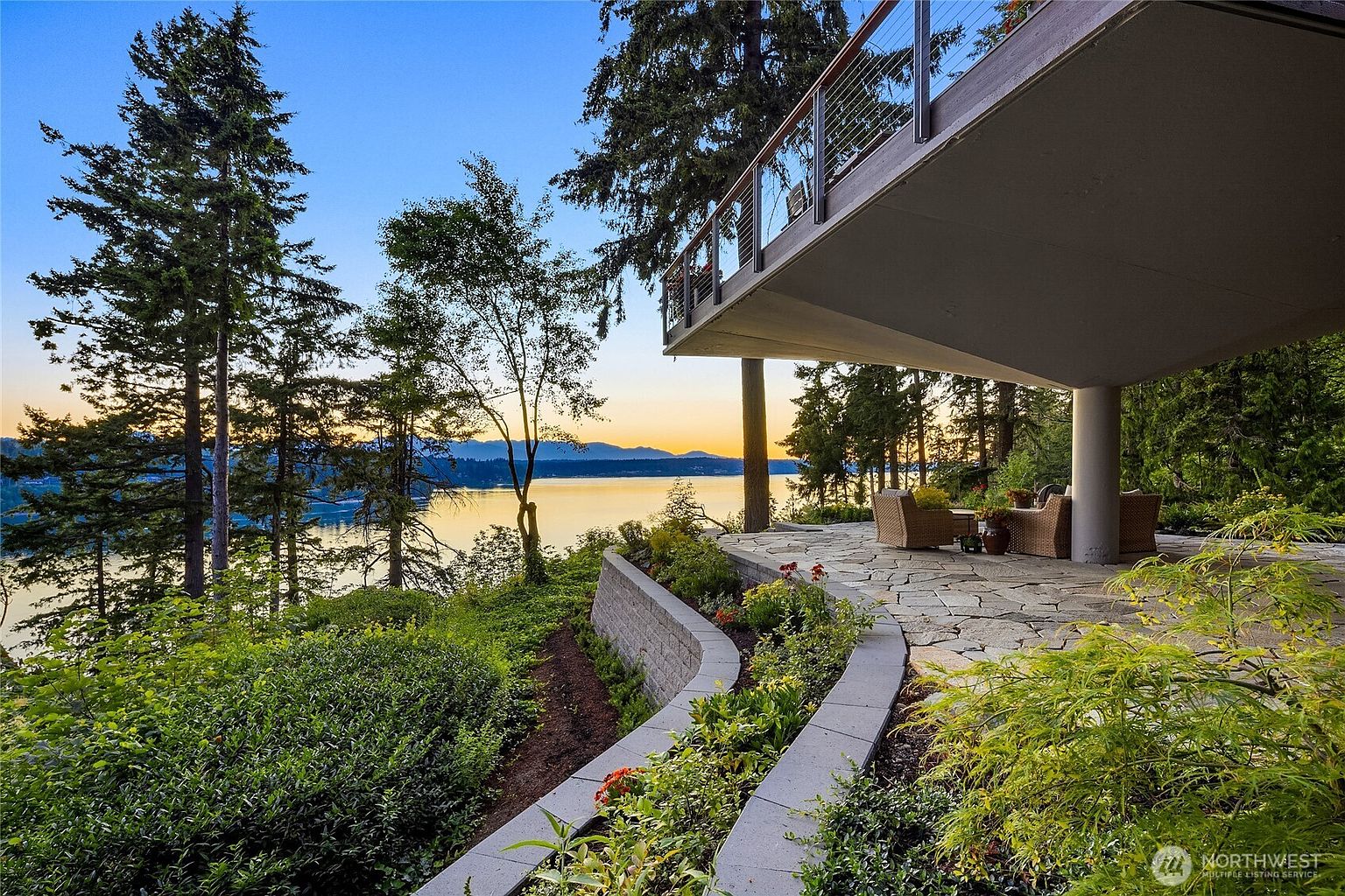 This image showcases a stunning outdoor patio and balcony area with breathtaking water views. The patio features elegant stone flooring and comfortable wicker furniture, creating an inviting space for relaxation and entertainment. A beautifully landscaped garden with retaining walls adds to the property's appeal, while the balcony offers a modern touch with its glass railings and sleek design, all overlooking a serene lake at sunset.