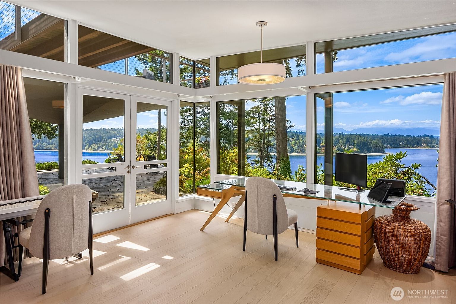 This is a bright and airy office space with floor-to-ceiling windows offering stunning water views. The room features a glass-top desk with wooden drawers, a modern chair, and a decorative woven vase. The light wood flooring and minimalist design create a serene and productive atmosphere.