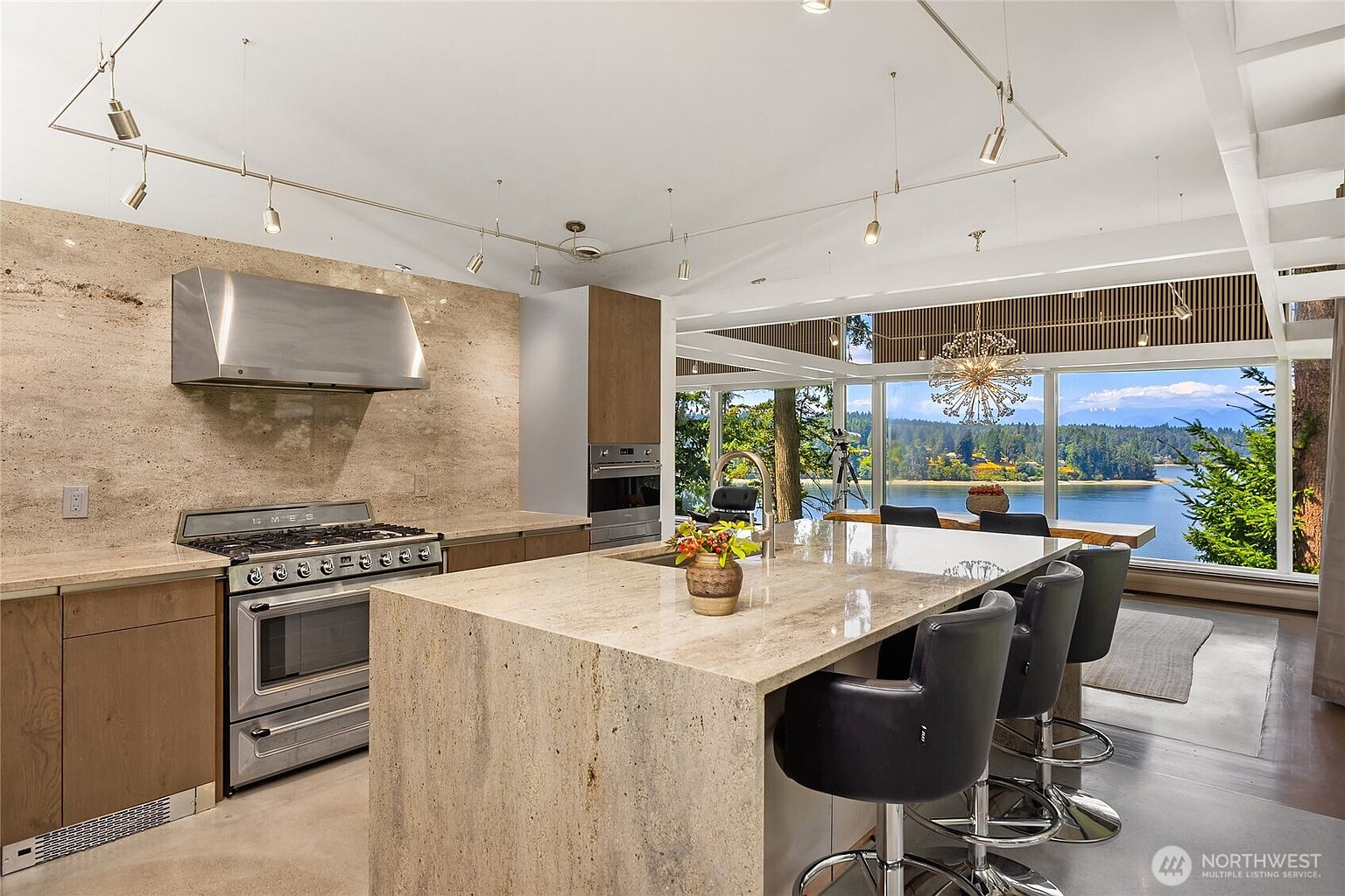 This is a bright and modern kitchen with a large island featuring bar seating and a sink. The kitchen boasts stainless steel appliances, including a range and hood, and a stone backsplash that complements the countertops. Large windows offer a stunning view of the water, enhancing the kitchen's appeal.