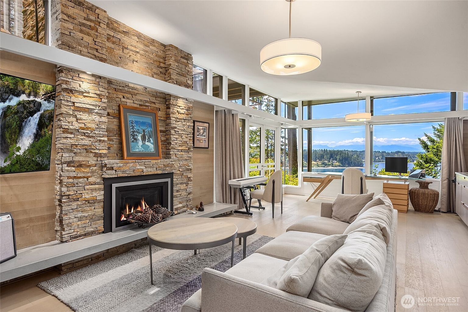 This is a bright and airy living room featuring a stone fireplace with a mounted television and artwork. Large windows offer stunning views of the water and surrounding landscape, complemented by neutral-toned furniture and modern lighting fixtures. The space exudes a sense of luxury and relaxation, perfect for entertaining or quiet enjoyment.