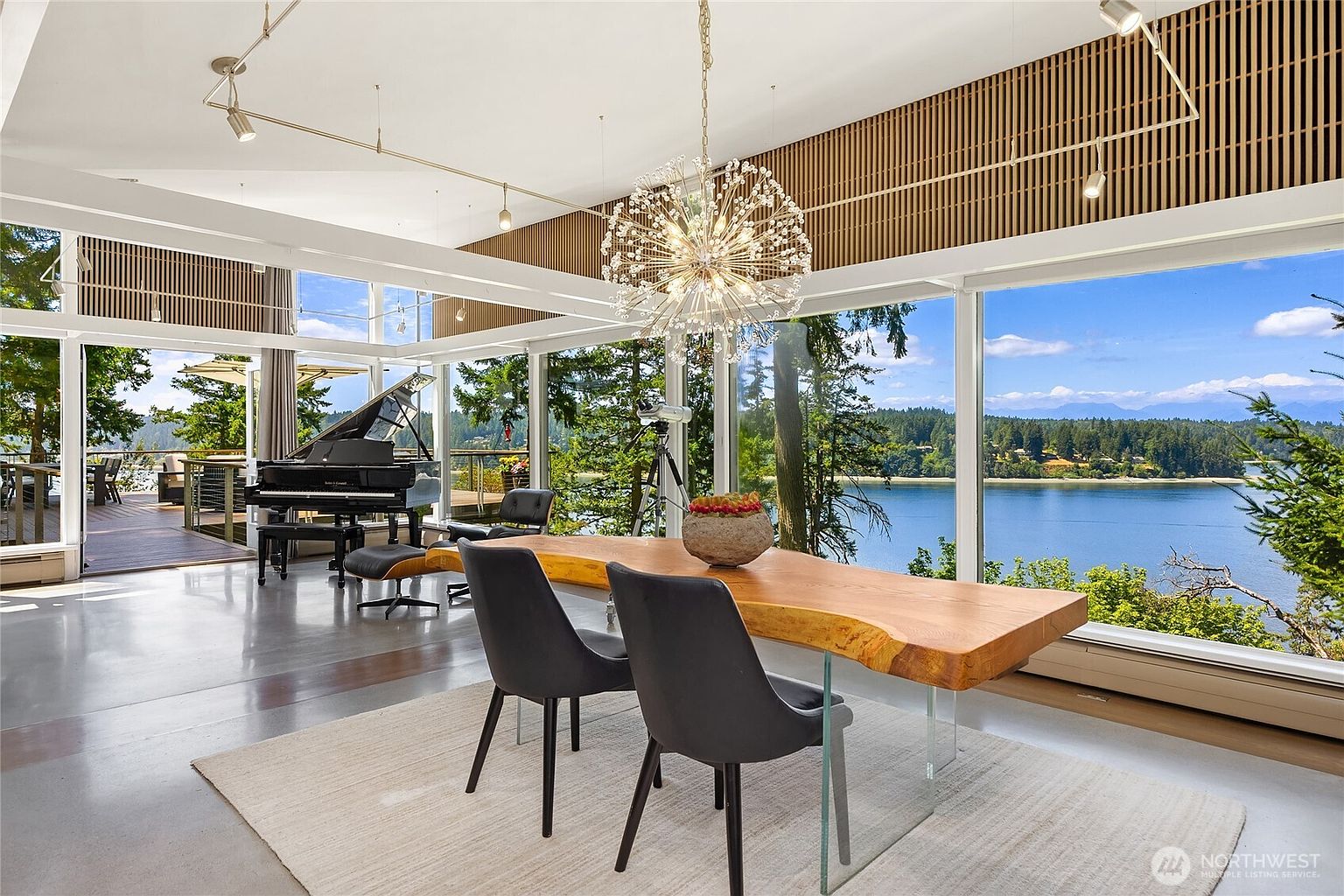 This is a bright and airy dining room featuring a unique, live-edge wooden table with clear acrylic legs, surrounded by modern black chairs. A grand piano sits in the background near a large window overlooking a waterfront view, and a striking chandelier hangs above the table. The room's design emphasizes natural light and a connection to the surrounding landscape.
