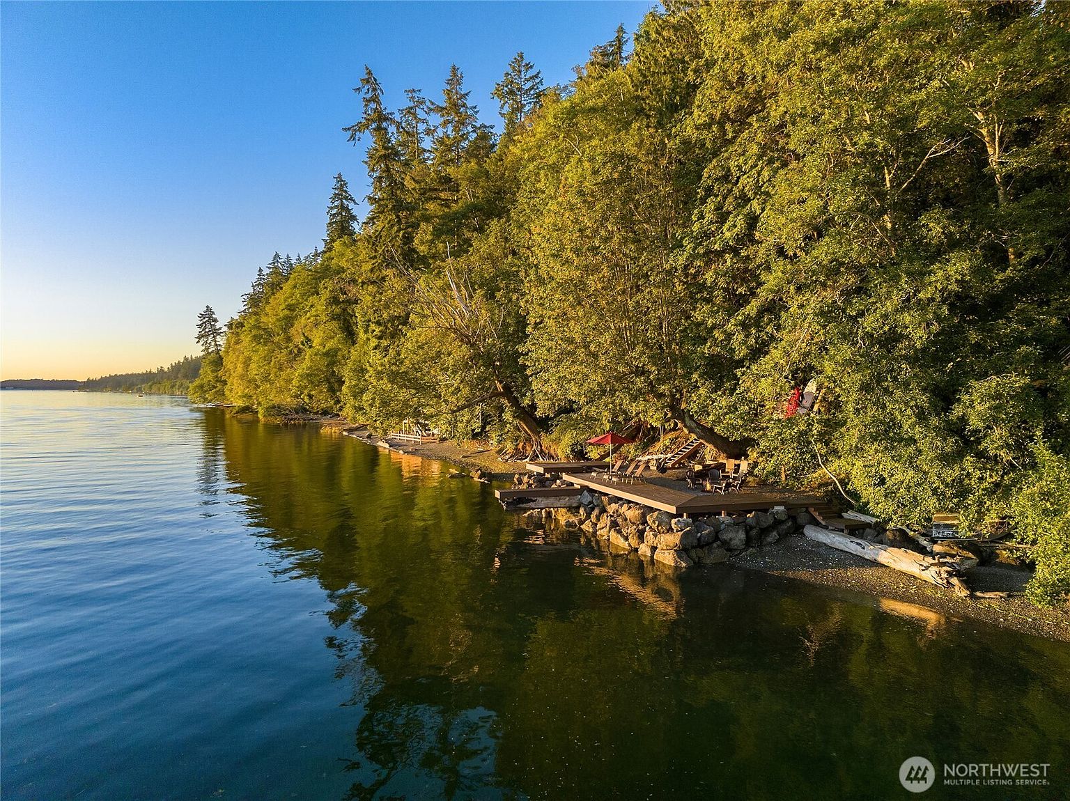 This exterior shot showcases a serene waterfront property with a private deck extending over the water. Lush greenery covers the hillside, providing a natural backdrop. The calm water reflects the trees and sky, creating a peaceful and inviting atmosphere, perfect for relaxation and enjoying the natural surroundings.