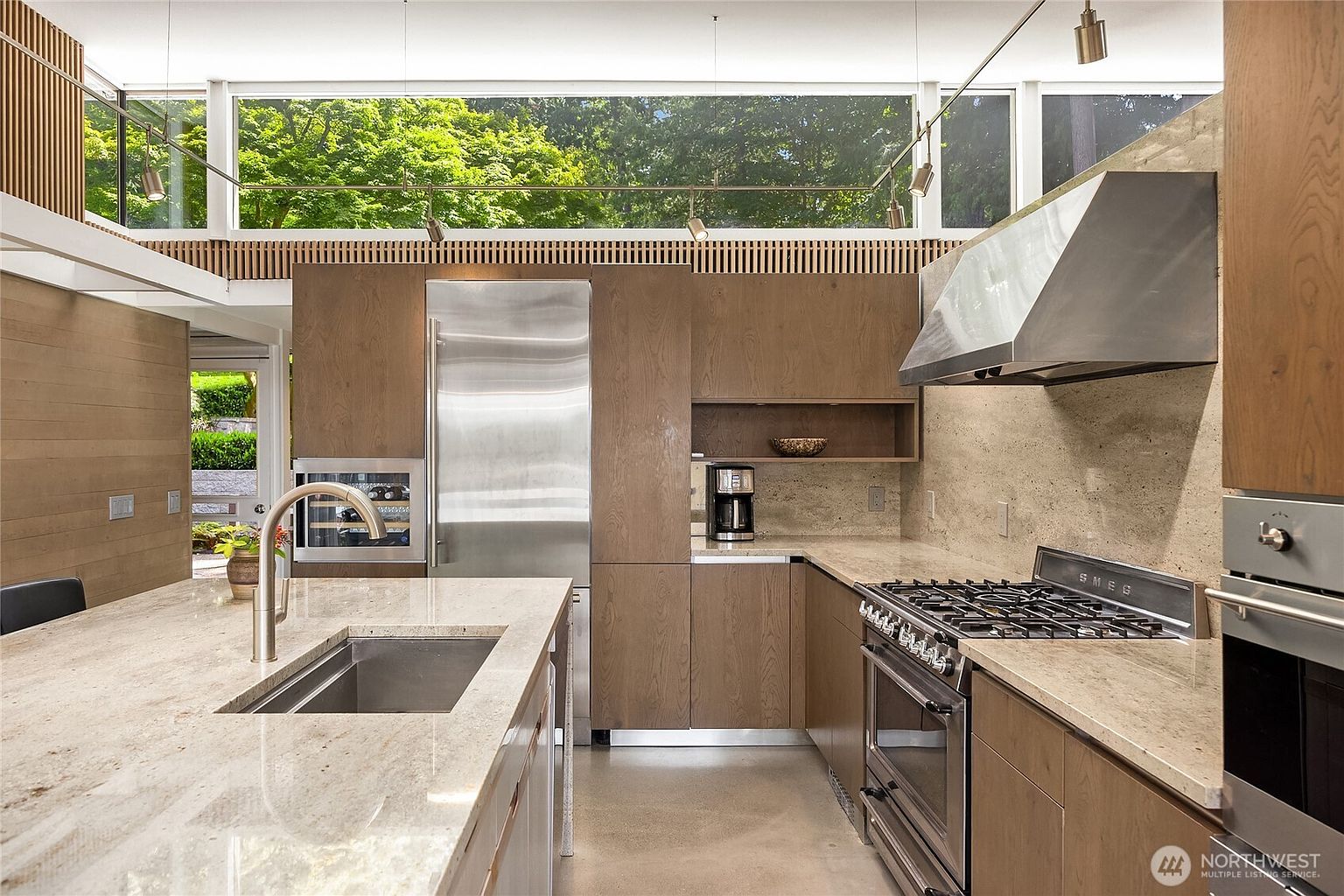 This is a modern kitchen featuring sleek, wood-paneled cabinetry and stainless steel appliances. A large island with a light-colored countertop dominates the foreground, while a stainless steel refrigerator and a gas range are visible in the background. The kitchen is well-lit with natural light streaming through a large window, offering a view of lush greenery outside.