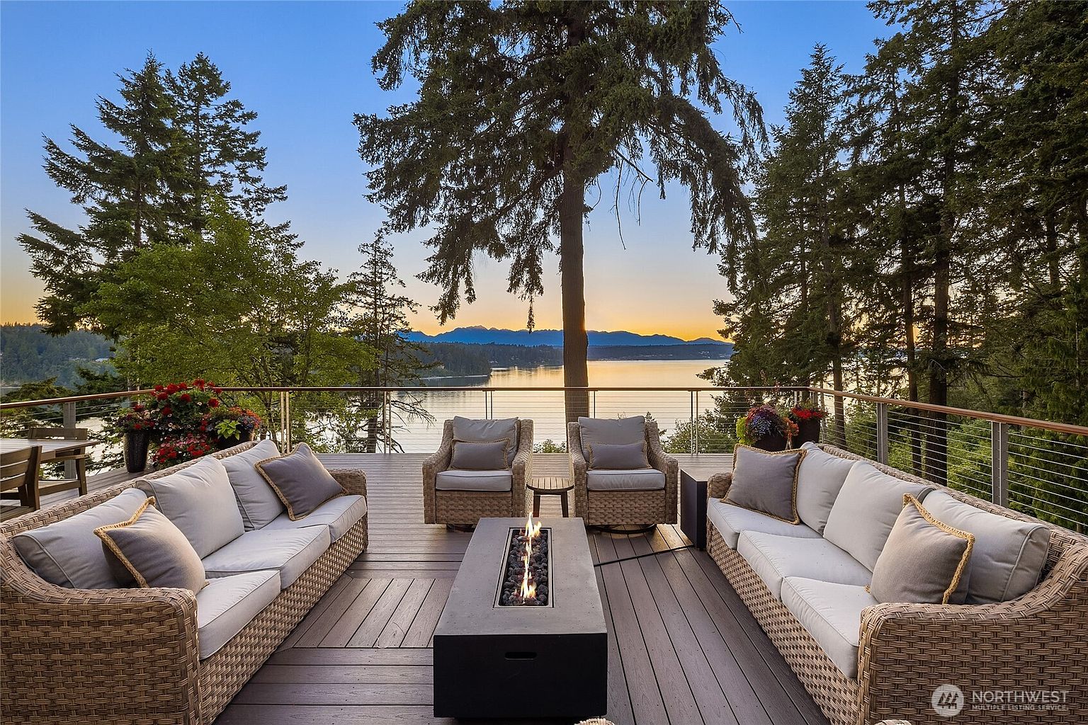This image showcases a luxurious outdoor deck area, featuring comfortable wicker sofas and armchairs arranged around a modern fire pit. The deck offers stunning views of a serene lake and distant mountains, framed by lush trees. The setting suggests a perfect space for relaxation and entertaining, highlighting the property's outdoor living potential.