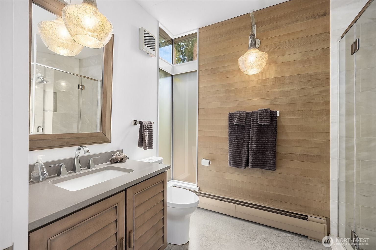 This is a well-lit bathroom featuring a modern design with a wood accent wall and a floating vanity. The vanity has a light-colored countertop and wooden cabinets with horizontal slats. A large mirror hangs above the sink, and unique pendant lighting adds a touch of elegance. The bathroom also includes a toilet and a glass-enclosed shower, creating a clean and functional space.