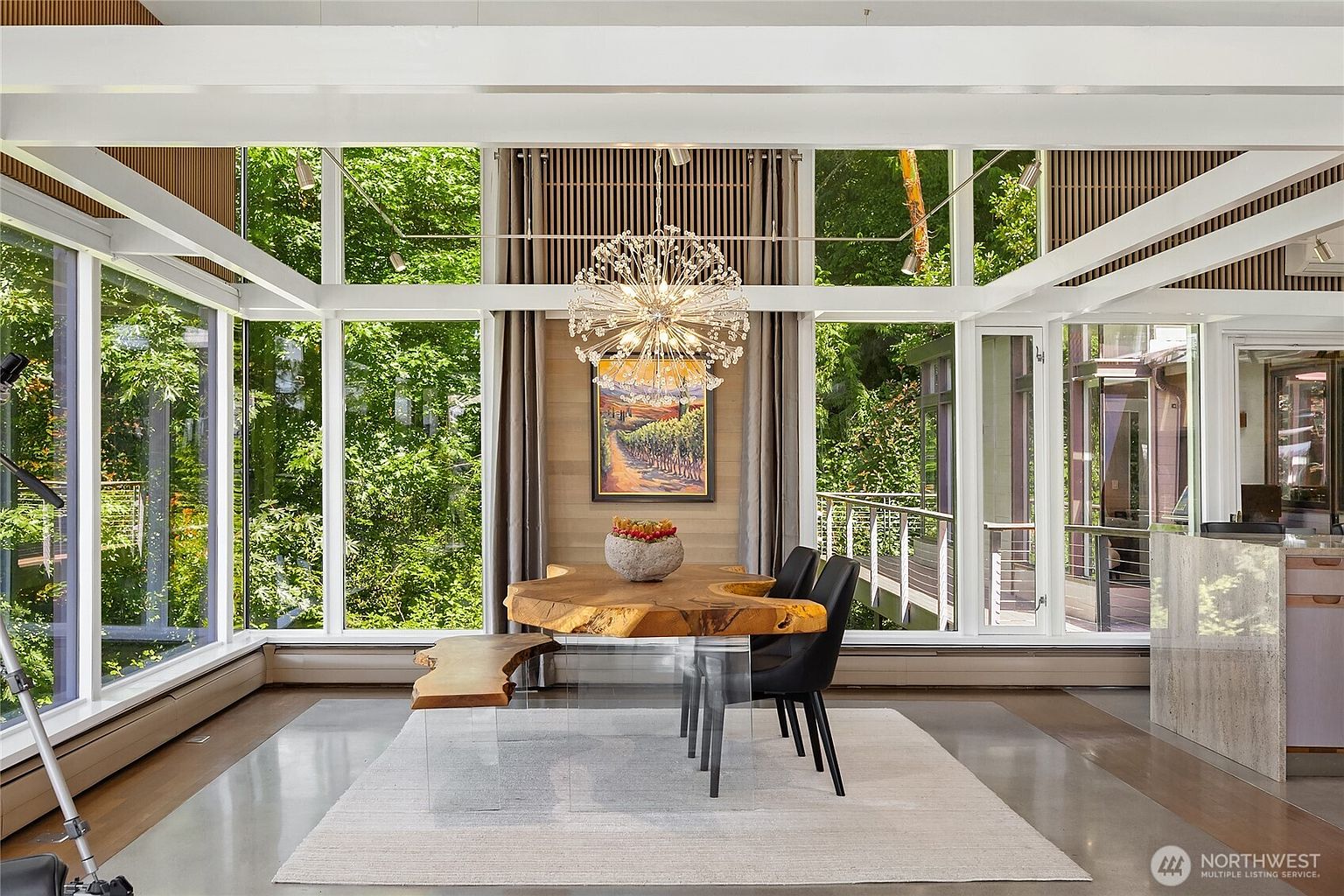 This is a bright and airy dining room featuring a unique live-edge wood table with a glass base, surrounded by modern black chairs. A striking chandelier hangs above the table, and a large painting adorns the wall. The room is enclosed by expansive windows offering views of lush greenery, creating a seamless connection with the outdoors.