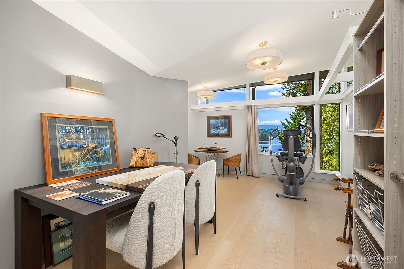 This interior shot showcases a bright and airy office space with a modern aesthetic. The room features a dark wood desk with neutral-toned chairs, a bookshelf, and large windows offering a scenic view of the water. An elliptical machine suggests a dual-purpose space for work and exercise.