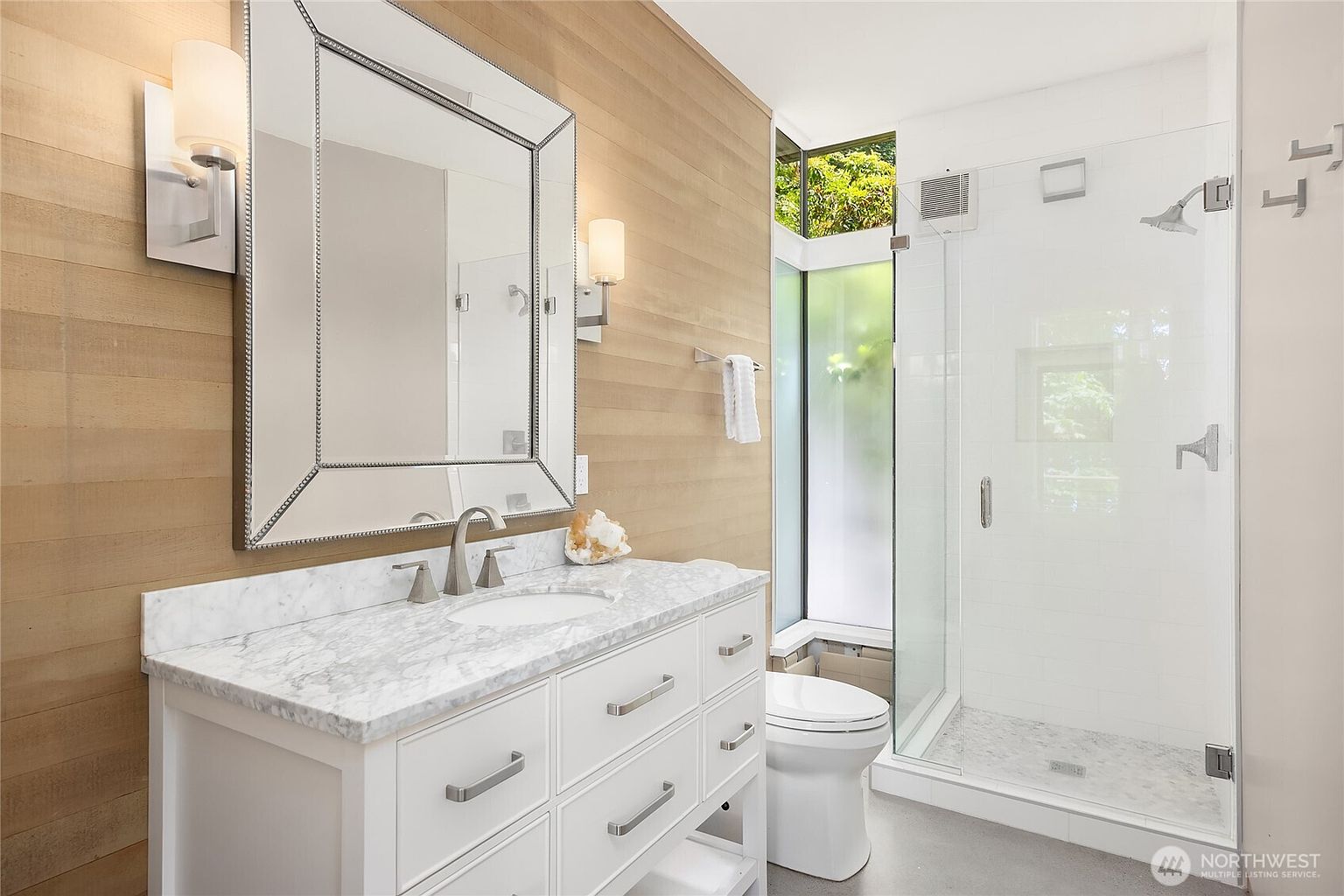 This is a well-lit primary bathroom featuring a white vanity with a marble countertop, a large mirror, and modern lighting fixtures. The bathroom includes a glass-enclosed shower and a toilet, with a window providing natural light and a view of greenery. The overall design is clean and contemporary, creating a spa-like atmosphere.