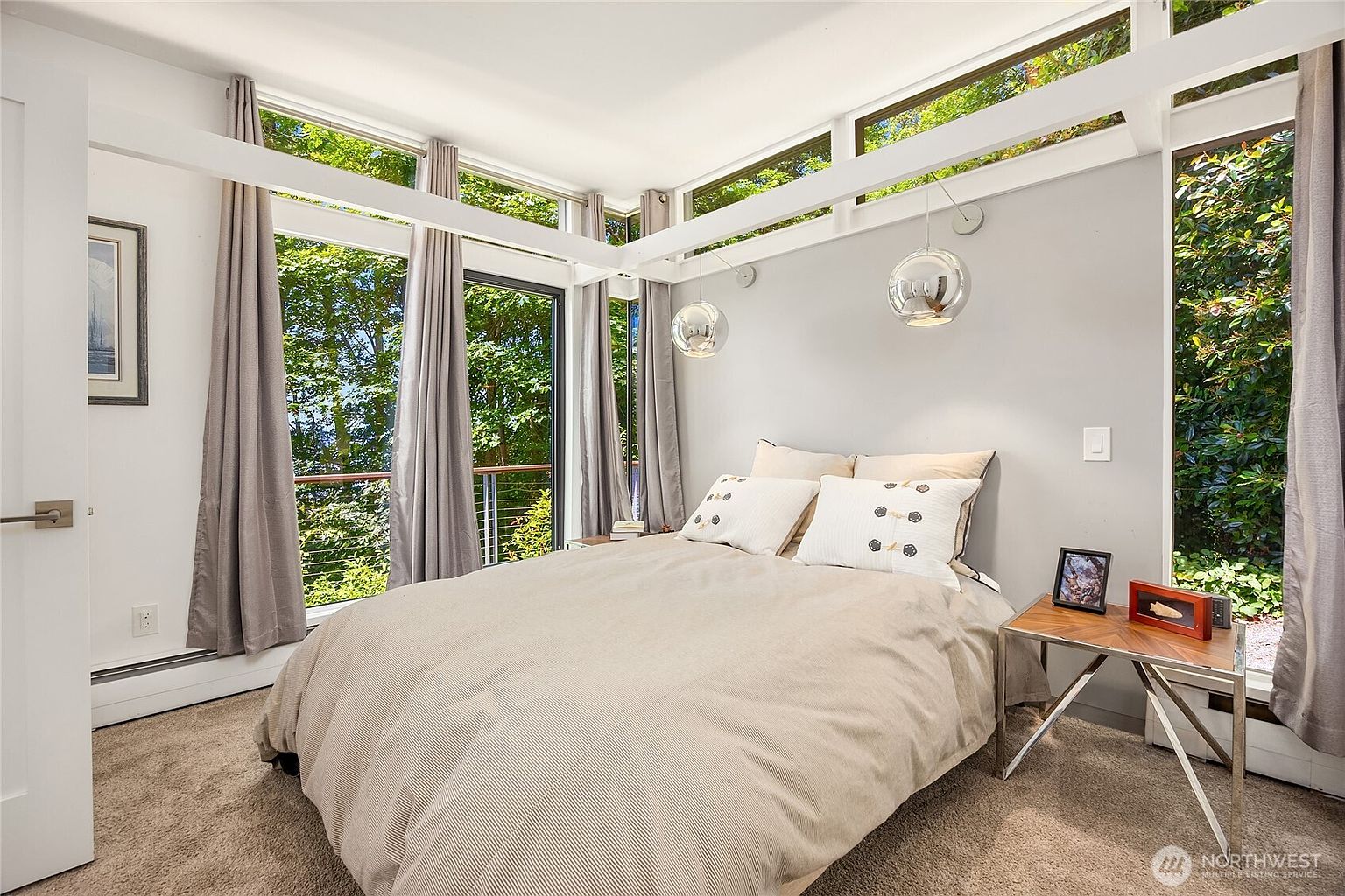 This is a bright and airy primary bedroom featuring large windows that offer a view of lush greenery. The room is decorated in neutral tones, with a comfortable bed adorned with decorative pillows. Modern globe pendant lights hang from the ceiling, adding a touch of elegance to the space.