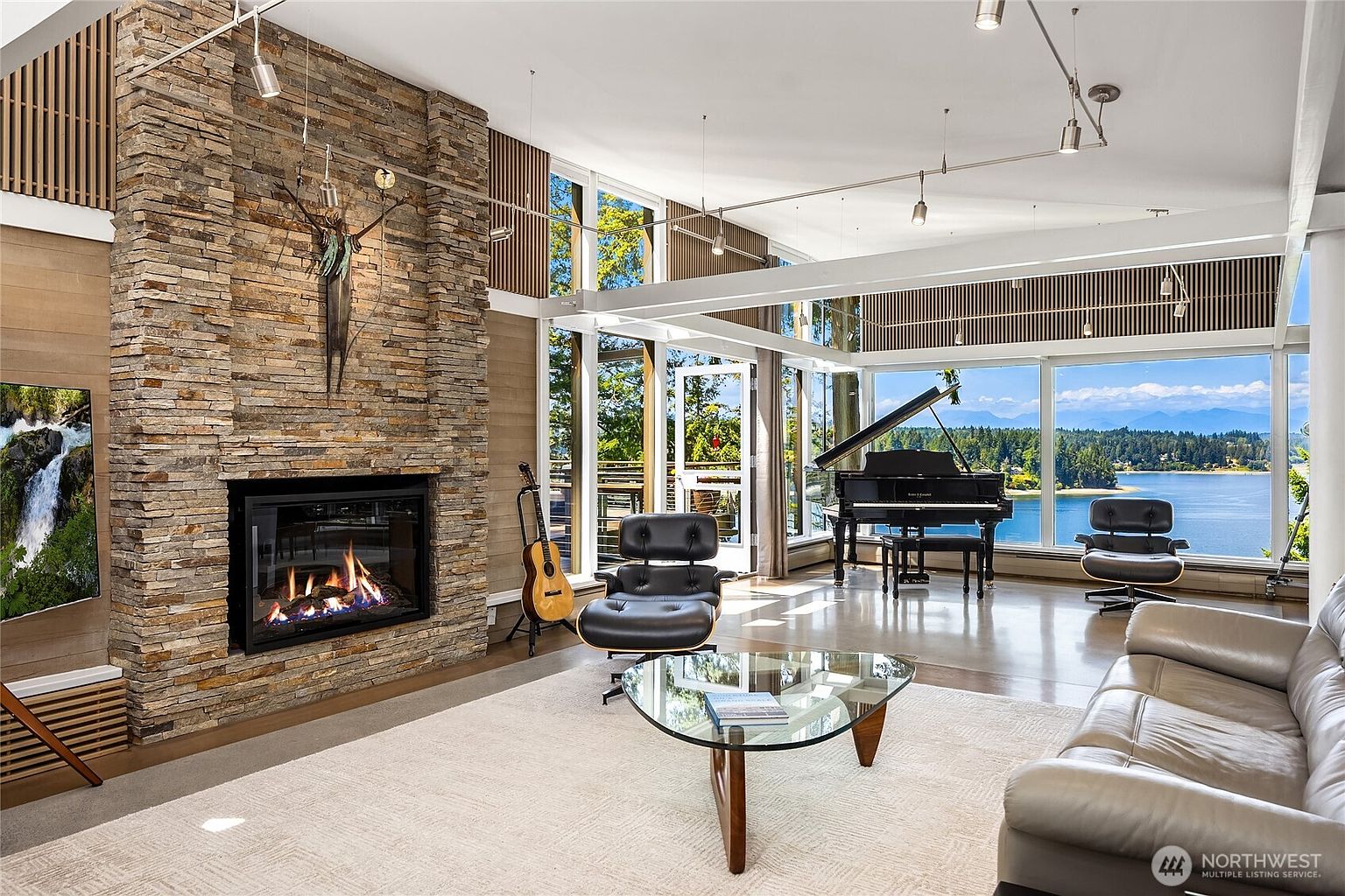 This is a spacious living room featuring a stone fireplace, a grand piano, and large windows with a scenic water view. The room is furnished with modern furniture, including a leather sofa and a glass coffee table. The overall style is contemporary and luxurious, emphasizing natural light and open space.