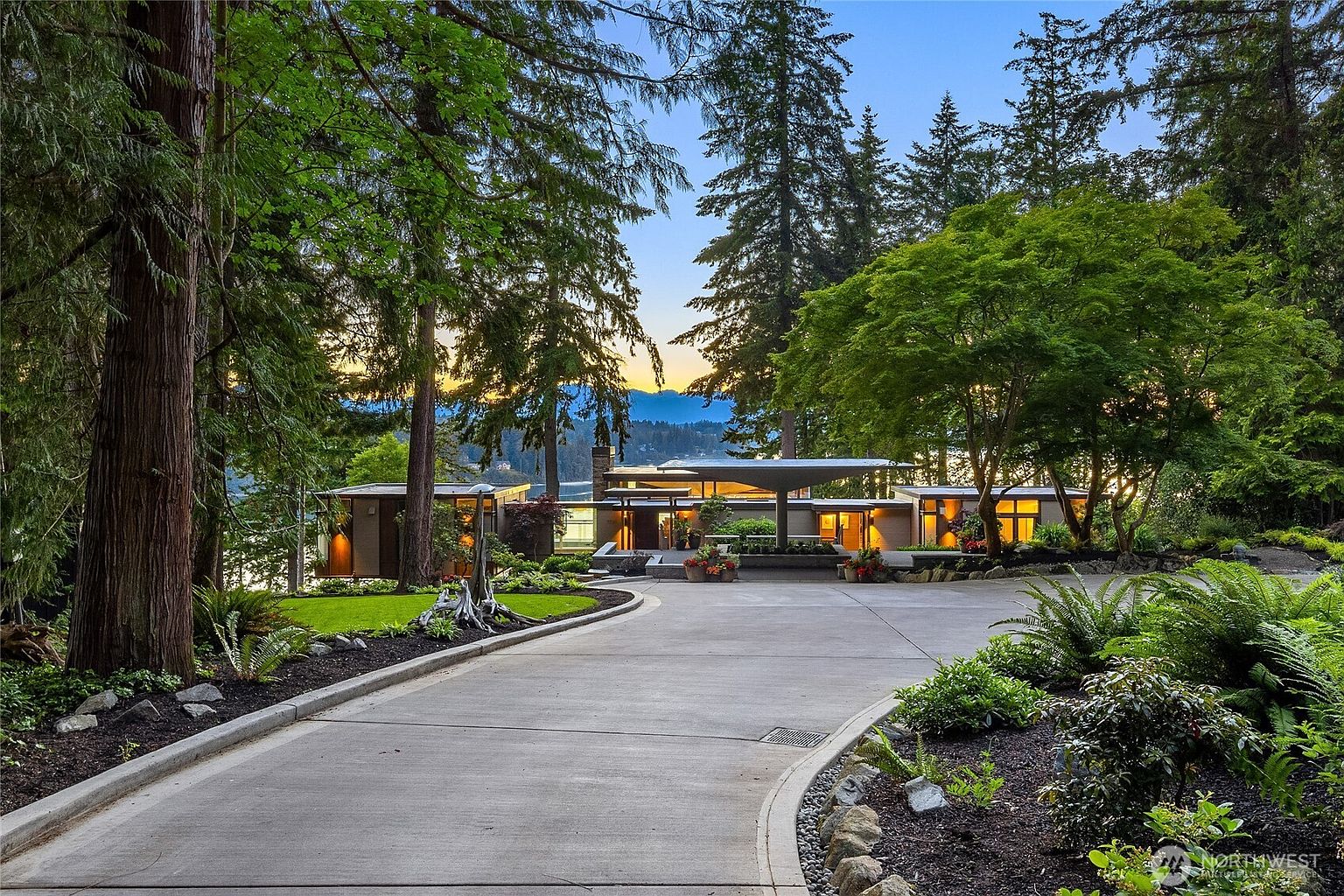 The image showcases the front exterior of a modern, single-story home nestled among mature trees. A long, curved driveway leads to the entrance, flanked by meticulously landscaped gardens. The home features a flat roof with cantilevered elements, large windows, and warm interior lighting, creating an inviting and sophisticated ambiance.
