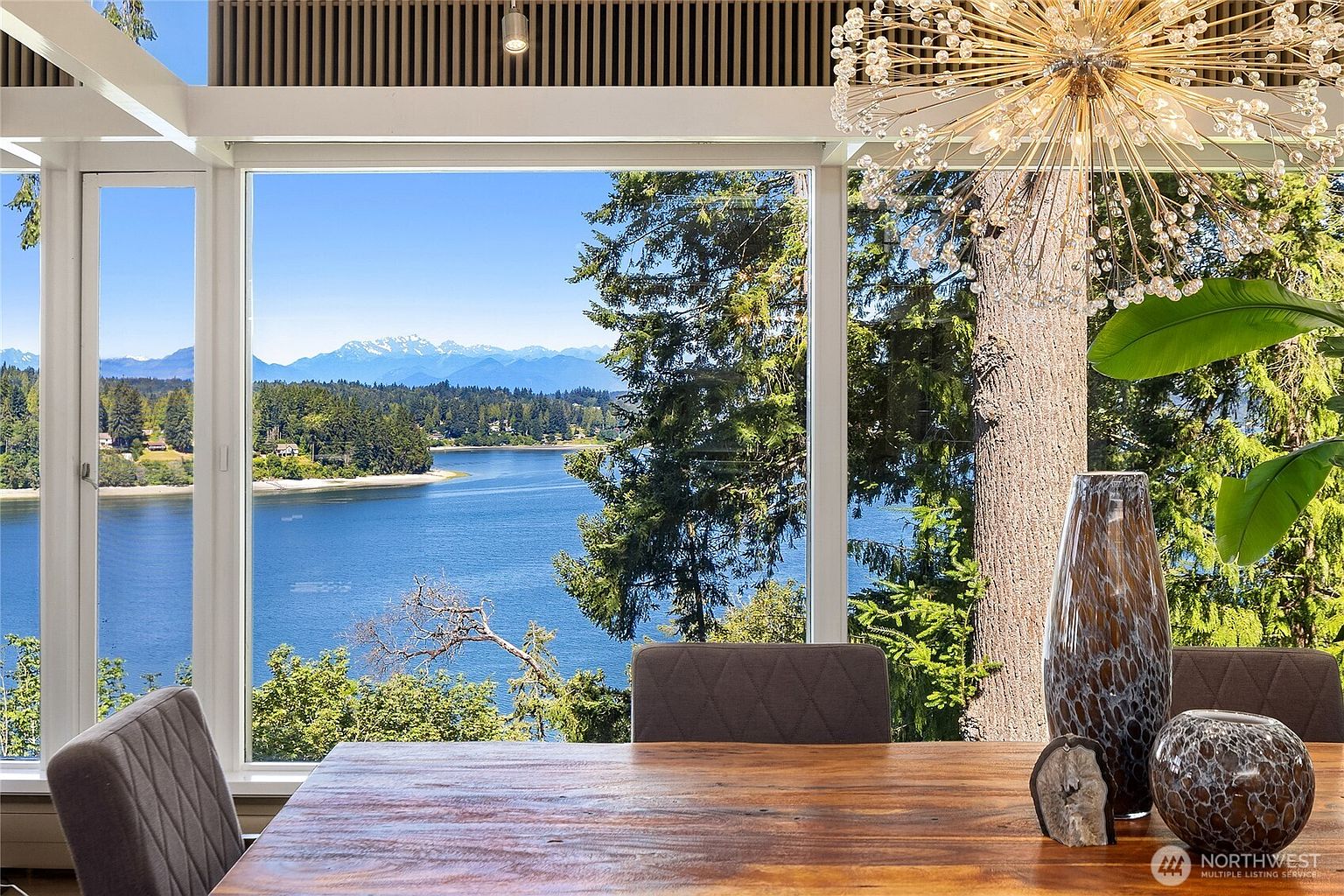 This image showcases a dining room with a stunning view of a lake and mountains through large windows. The room features a wooden dining table with chairs, decorative vases, and a modern chandelier. The overall impression is one of elegance and tranquility, highlighting the property's desirable location and sophisticated interior design.