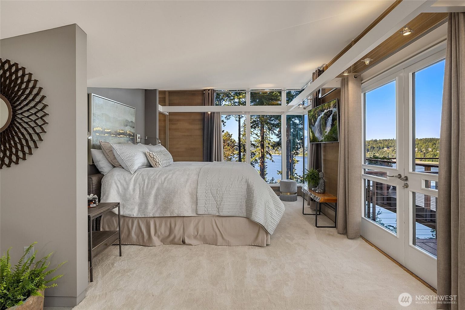 This is a luxurious primary bedroom with a stunning view of the lake. The room features a large bed with neutral bedding, floor-to-ceiling windows, and access to a balcony. The decor is modern and elegant, creating a serene and inviting atmosphere.
