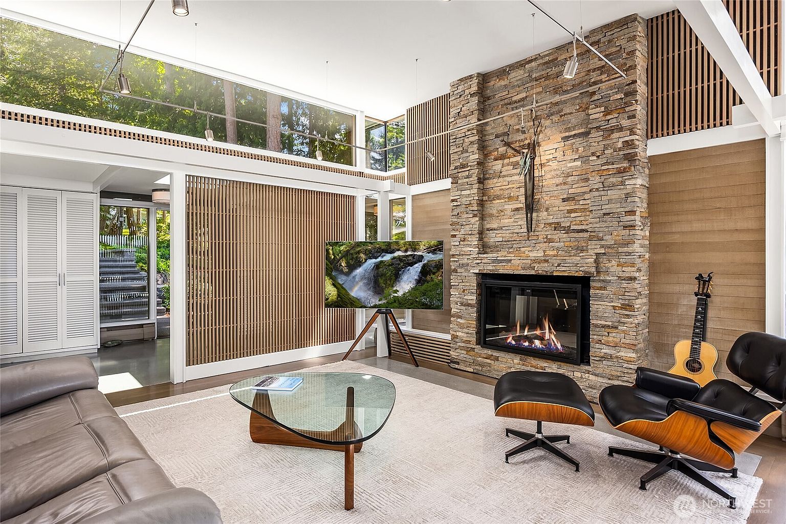 This is a stylish living room featuring a stone fireplace, a modern TV stand, and a glass coffee table. The room is well-lit with natural light and features a neutral color palette with wood accents. The overall impression is modern and inviting, perfect for relaxation and entertainment.