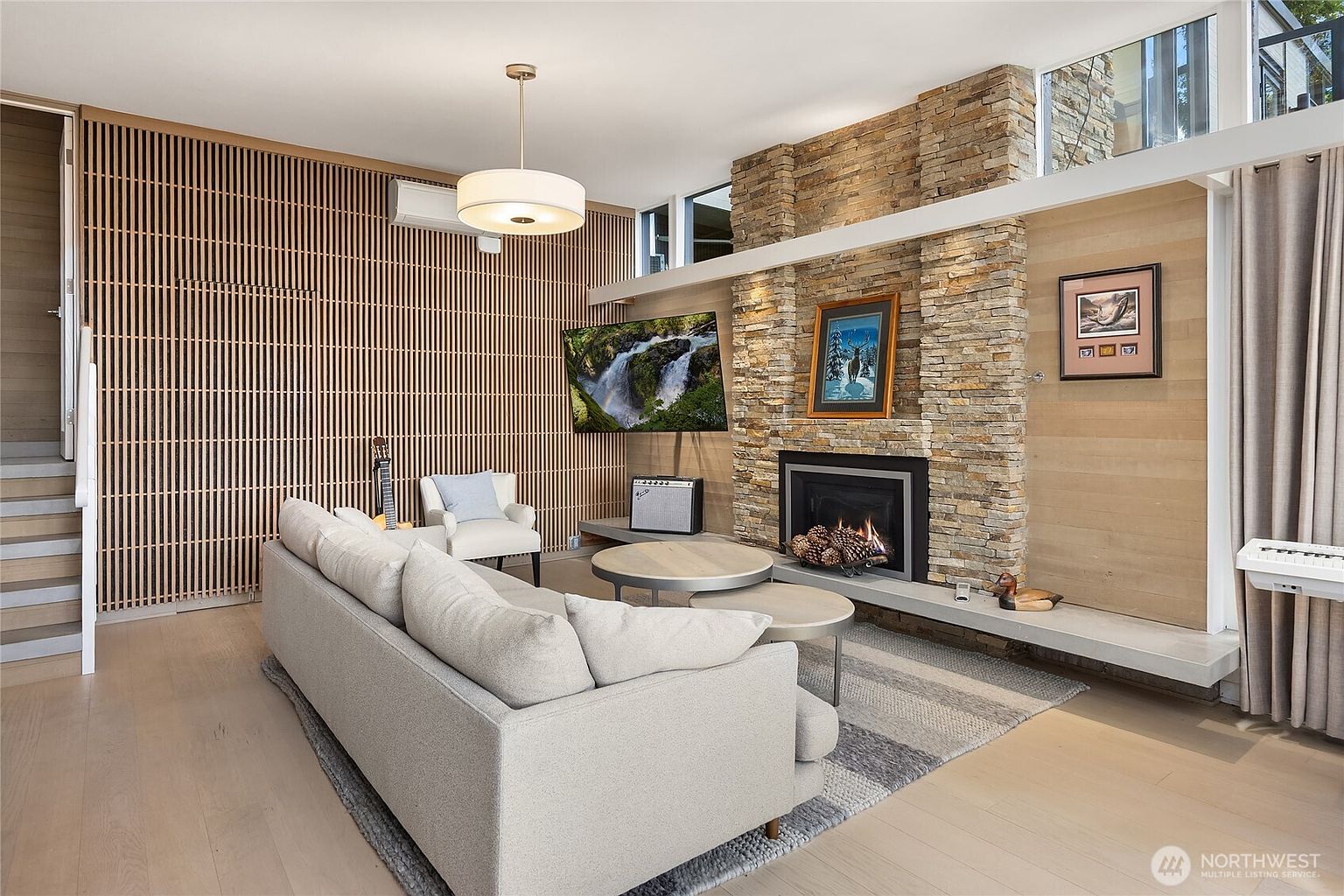 This is a warm and inviting living room featuring a stone fireplace, a comfortable sofa, and modern decor. The room is well-lit with natural light and a stylish pendant light fixture. The overall impression is one of comfort and contemporary elegance, perfect for relaxation and entertaining.