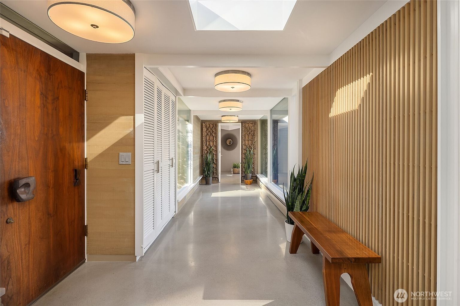 This is an interior shot of a modern hallway. The hallway features a polished concrete floor, wood slat wall paneling, and a series of white louvered closet doors. Natural light streams in through a skylight and windows, illuminating the space and highlighting the clean lines and minimalist design.