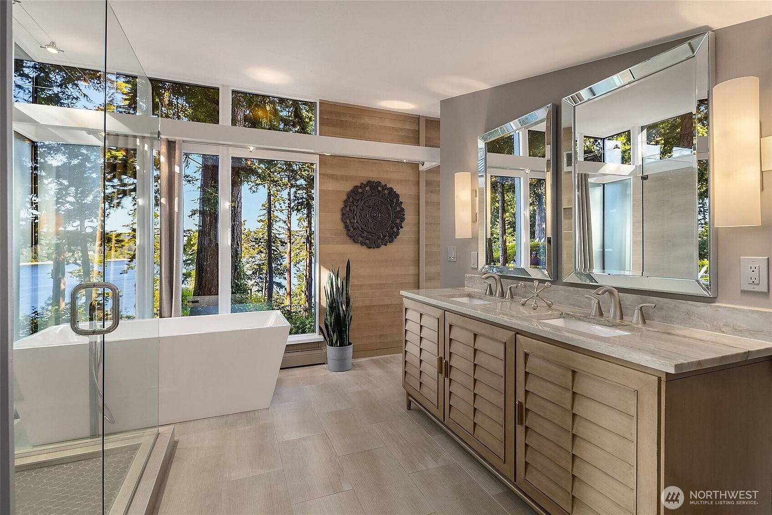 This is a luxurious primary bathroom featuring a freestanding soaking tub positioned in front of large windows with a lake view. The double vanity has a light wood finish with a marble countertop and modern fixtures. The room is well-lit with sconces and natural light, creating a spa-like atmosphere.