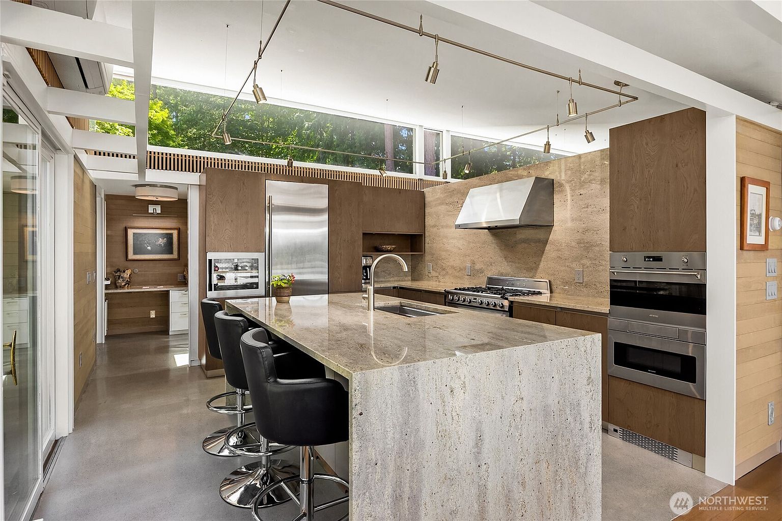 This is a modern kitchen with a large island featuring a stone countertop and bar seating. Stainless steel appliances, including a refrigerator and built-in ovens, complement the wood cabinetry. A long window above the cabinets provides natural light, and track lighting illuminates the space.
