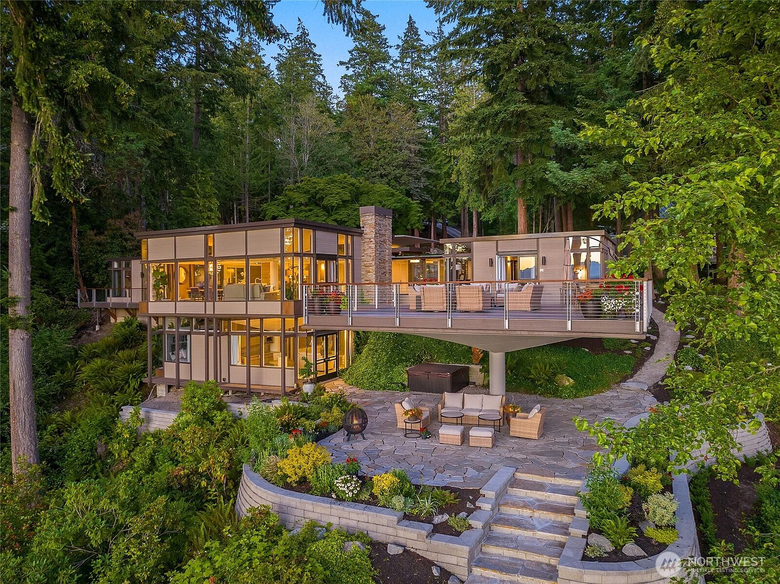 This aerial view showcases a modern, multi-level home nestled among lush trees. The house features large windows, a stone chimney, and an expansive deck with outdoor seating. A lower patio area includes a fire pit and additional seating, all surrounded by meticulously landscaped gardens and stone pathways.