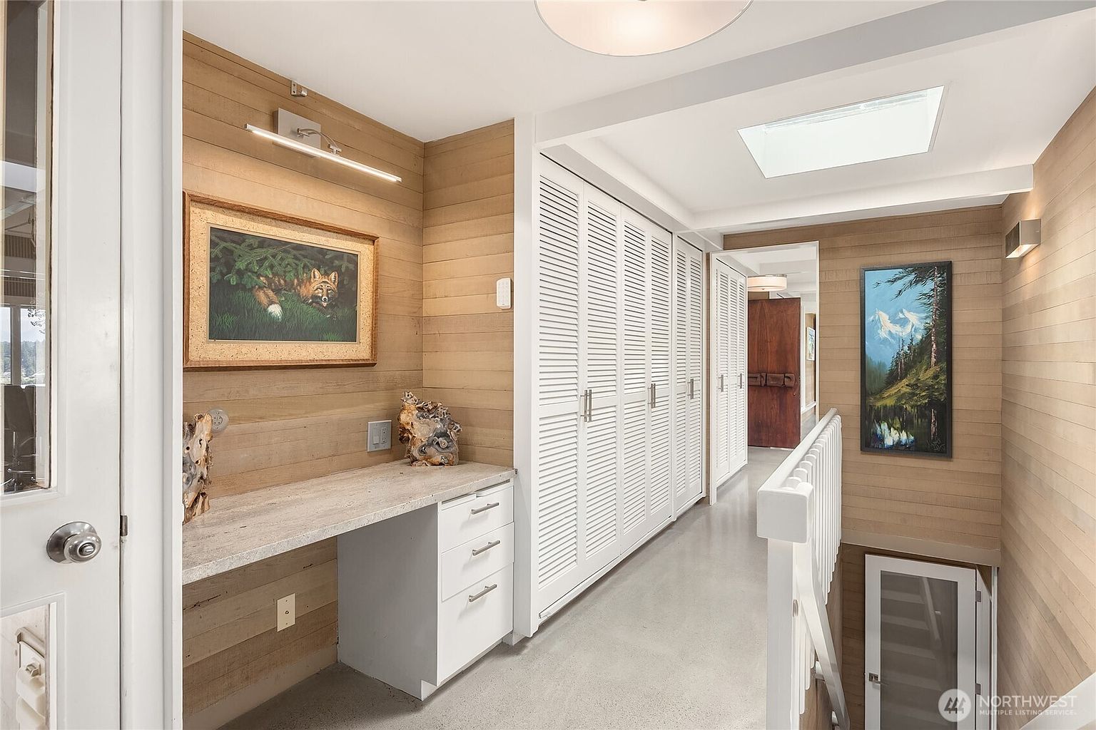 This interior shot showcases a well-lit hallway with wood-paneled walls and a series of white closet doors. A built-in desk area with drawers is visible on the left, adorned with artwork and decorative pieces. The hallway leads to a staircase and another room, creating a sense of depth and spaciousness.