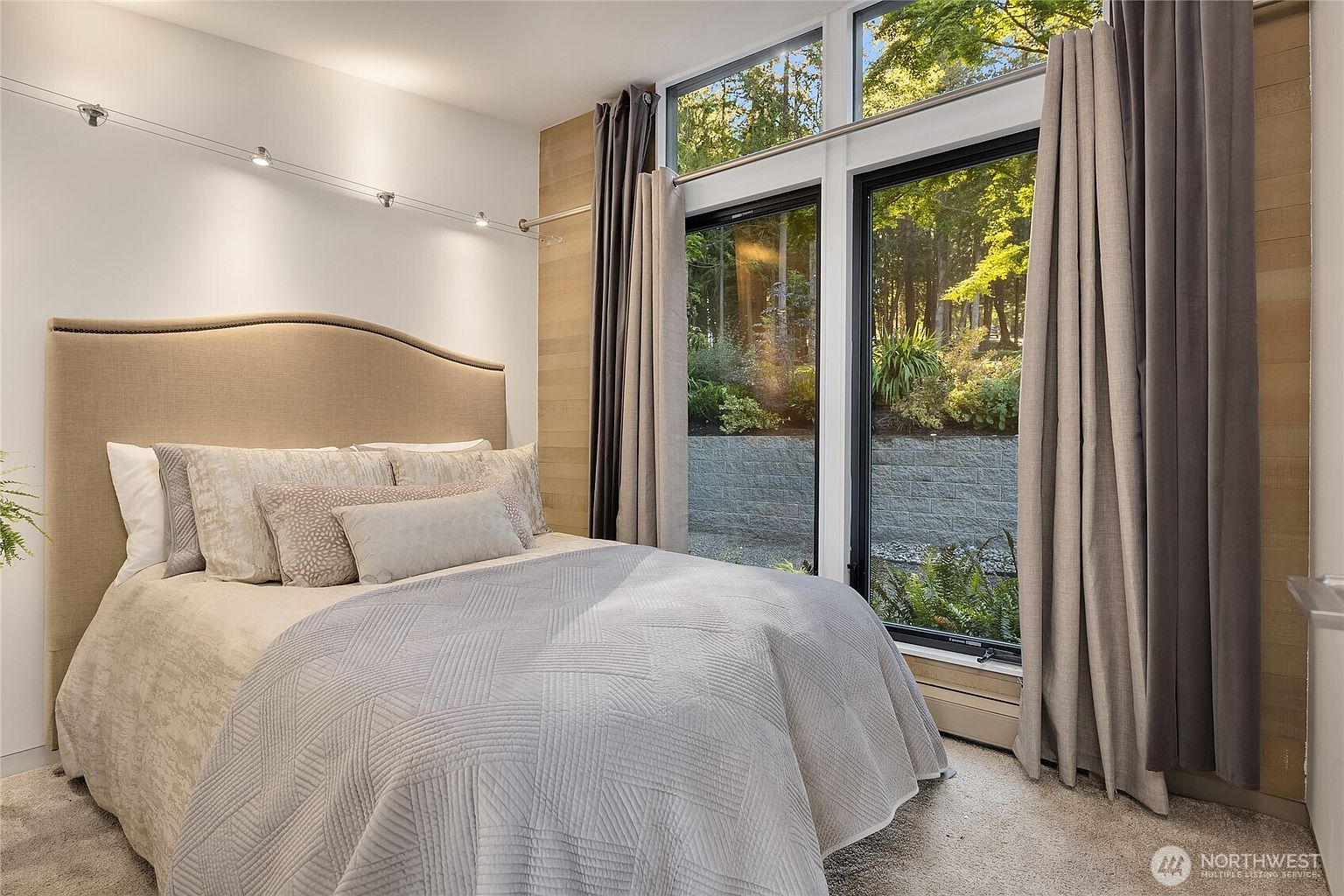 This is a well-lit primary bedroom featuring a large window with a view of a lush garden. The room is furnished with a bed that has a neutral-toned headboard and a light-colored quilt. Gray curtains frame the window, adding a touch of elegance and privacy to the space.