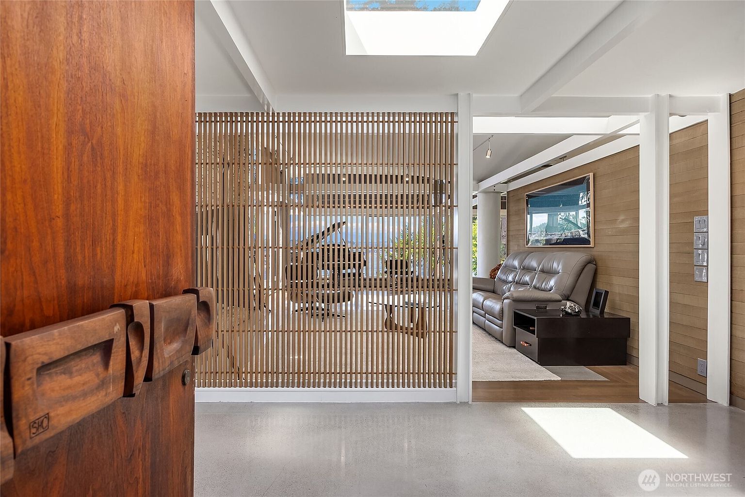 The image showcases an interior entryway featuring a striking wooden door with unique handle detailing. A slatted wooden screen partially obscures the view into a living area with a gray leather sofa and modern furnishings. The space is well-lit with natural light streaming through a skylight, and the flooring appears to be polished concrete, contributing to a contemporary aesthetic.