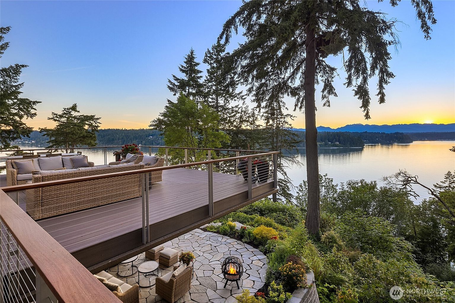 This image showcases a luxurious outdoor living space with a multi-level patio and deck overlooking a serene lake. The upper deck features comfortable wicker seating, while the lower patio includes a fire pit and additional seating, creating an inviting atmosphere for relaxation and entertainment. Lush landscaping surrounds the area, enhancing the natural beauty of the property.
