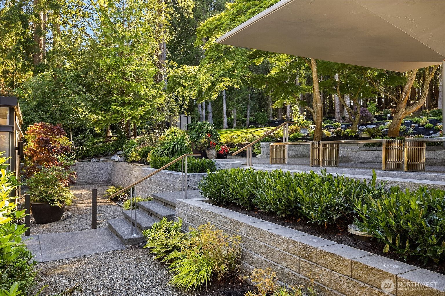This image showcases a beautifully landscaped yard and garden area. The design incorporates tiered levels with stone retaining walls, lush greenery, and mature trees, creating a serene and inviting outdoor space. A covered patio area with built-in seating provides an ideal spot for relaxation and entertainment.