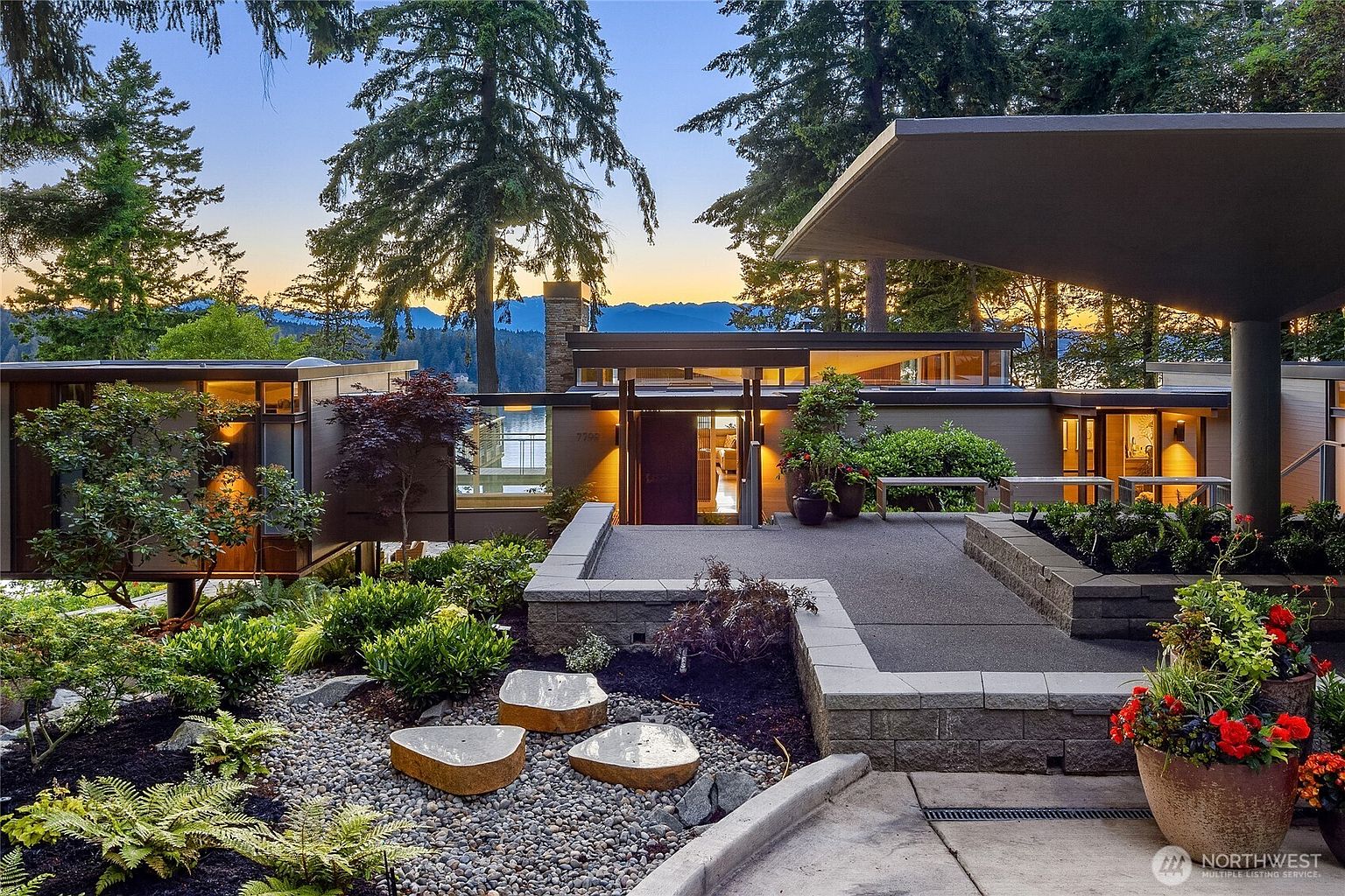 This is a front exterior view of a modern home with a unique architectural design. The house features a flat roof, large windows, and a stone facade. The landscaping includes a variety of plants, trees, and decorative rocks, creating a serene and inviting entrance. The overall impression is one of sophisticated elegance and natural beauty.
