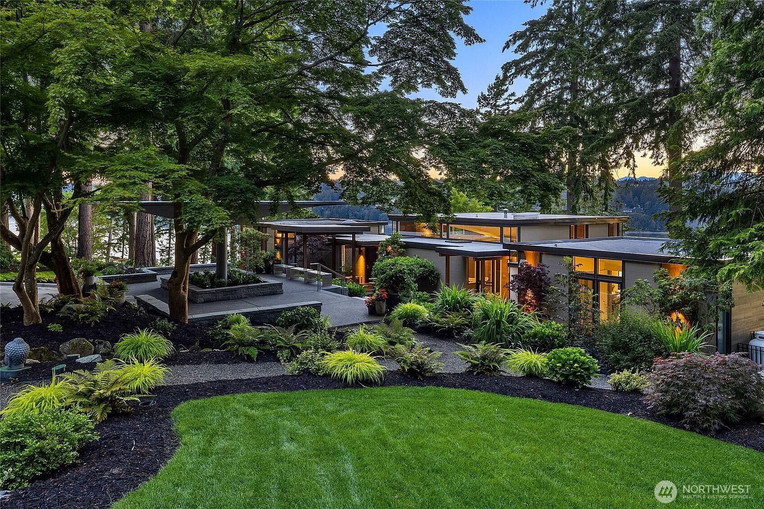 This image showcases a beautifully landscaped yard and garden area of a modern home. The lush green lawn is bordered by dark mulch and various plants, leading to a stone patio and the contemporary architecture of the house. The scene is framed by mature trees, creating a serene and private atmosphere, perfect for outdoor relaxation and entertaining.