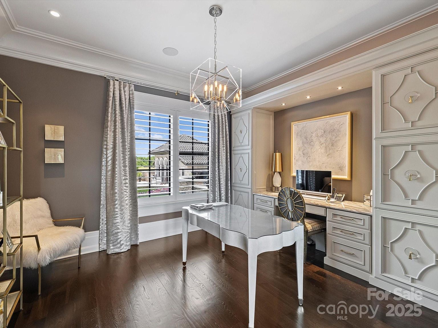 This elegant home office features dark hardwood flooring, a central desk with a sleek finish, and a plush, fur-covered accent chair. Built-in cabinetry with decorative paneling surrounds a desk area, which is illuminated by recessed lighting and a modern geometric chandelier. Large windows with shutters and metallic curtains provide natural light, creating a sophisticated and professional atmosphere.
