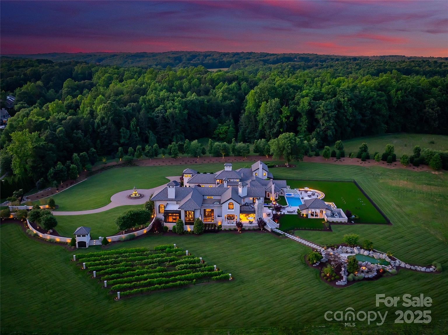This stunning aerial perspective captures a sprawling luxury estate at dusk, set against a backdrop of dense forest and a vibrant sunset. The property features a large multi-gabled residence with a swimming pool, a private vineyard, and a manicured putting green, all surrounded by expansive, well-maintained lawns. The warm glow from the interior lighting and outdoor fixtures creates an inviting and sophisticated atmosphere for this high-end residential estate.