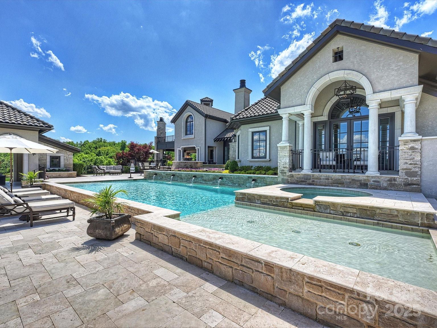 This luxurious outdoor living space features a multi-tiered swimming pool and spa complex set against a grand, light-toned stone residence. The scene includes a shallow tanning ledge, a raised spa, and a series of water features cascading into the main pool, all surrounded by expansive stone decking and lounge chairs. The architectural style is sophisticated and Mediterranean-inspired, offering a serene and high-end resort-like atmosphere.