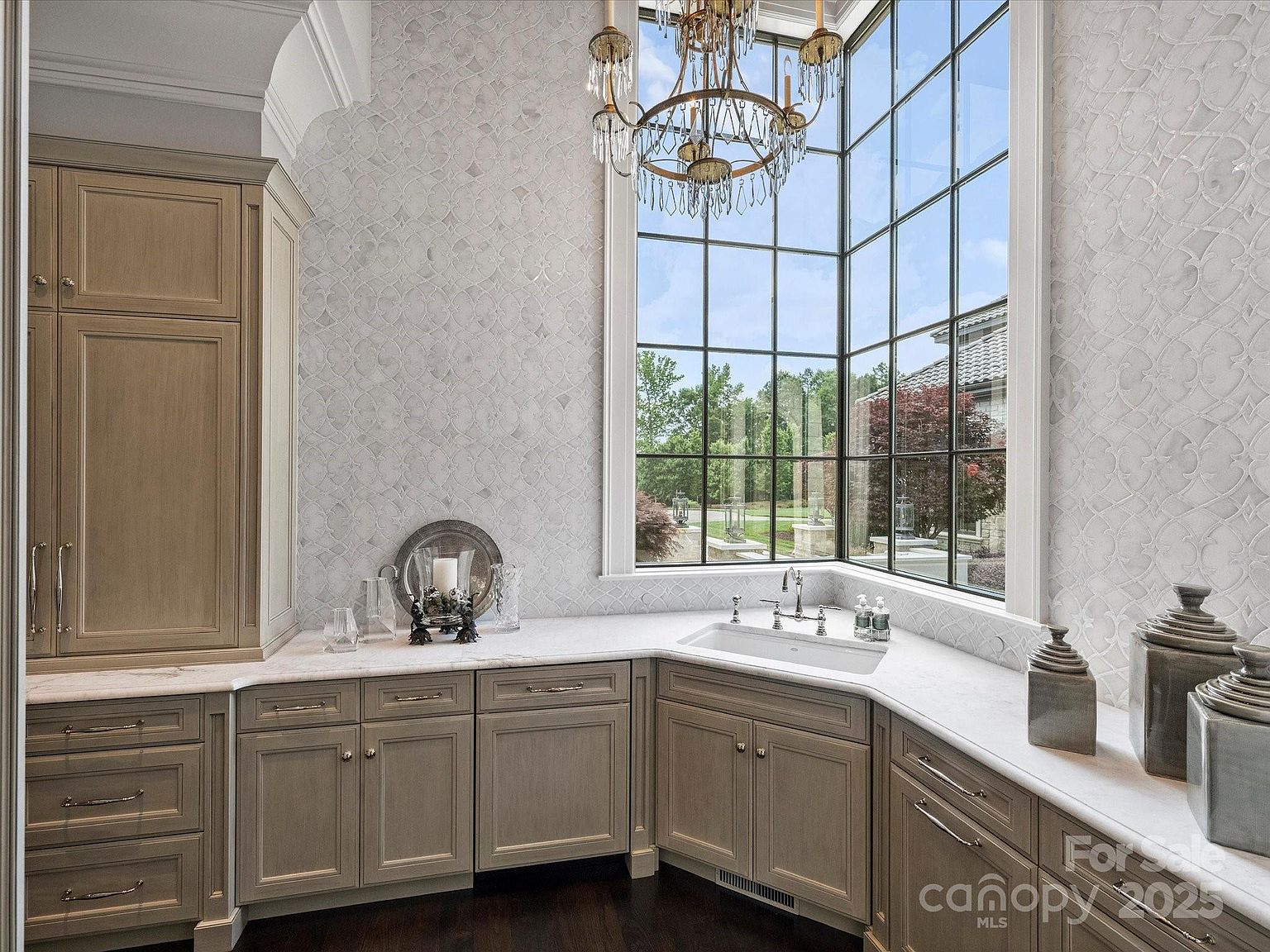 This elegant kitchen corner features light-toned cabinetry with classic hardware and a light-colored stone countertop. A large, multi-pane corner window arrangement provides ample natural light and a view of the outdoors, complemented by a decorative chandelier hanging above. The walls are adorned with a subtle, patterned tile backsplash, creating a sophisticated and bright atmosphere.