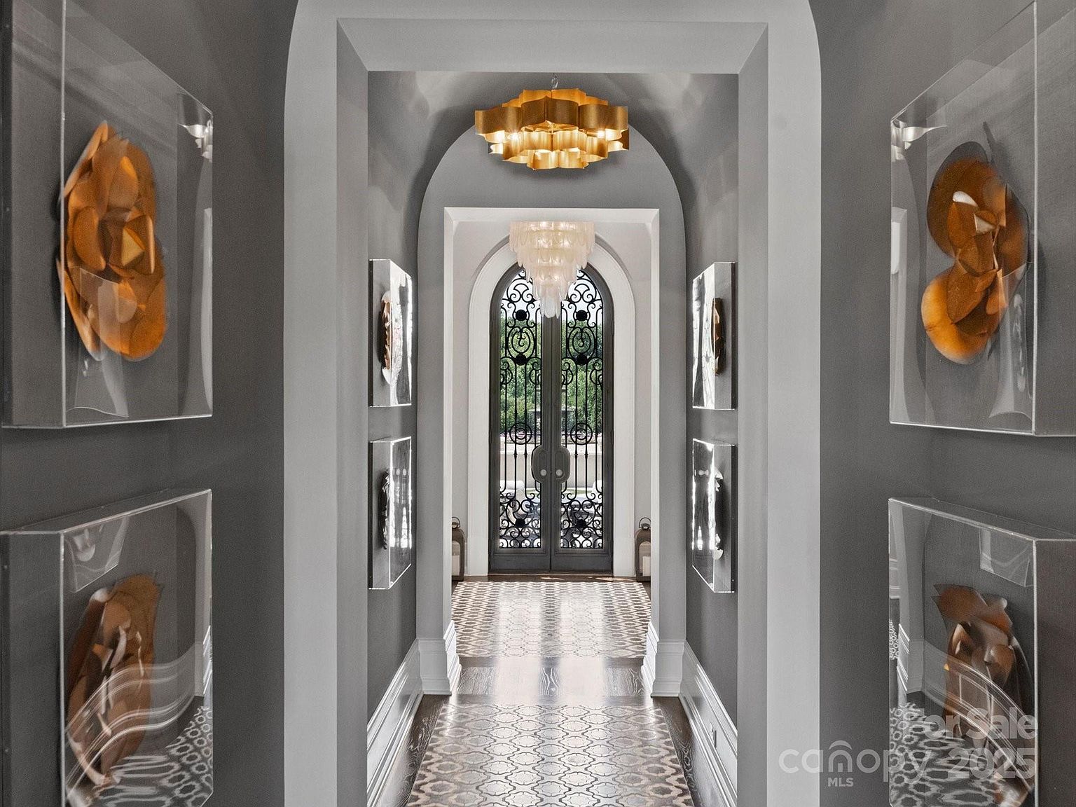 This elegant, symmetrical hallway features a vaulted ceiling with a gold-toned tiered chandelier and a secondary crystal chandelier near the arched iron-framed glass doors. The walls are painted a sophisticated dark gray, adorned with four framed abstract art pieces in shadow boxes. The floor is finished with a decorative patterned tile runner that leads the eye toward the bright, light-filled entryway.