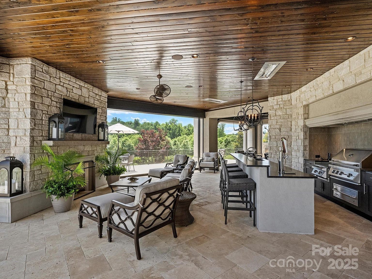 This expansive covered outdoor living space features a stone-clad chimney breast with an integrated television and a built-in outdoor kitchen equipped with a stainless steel grill. The area is furnished with comfortable lounge seating and a bar-style counter, all set against a backdrop of natural scenery visible through the open-air design. The space is finished with a wood-paneled ceiling and stone tile flooring, creating a sophisticated and inviting atmosphere for entertaining.