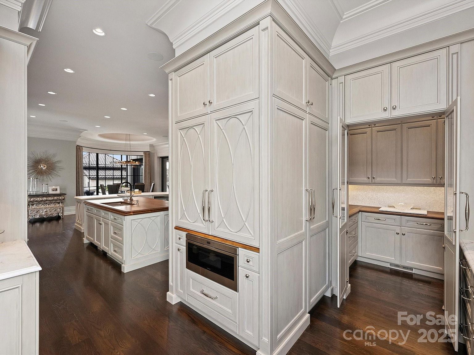 This sophisticated kitchen features custom light-toned cabinetry with intricate detailing and a central island topped with a rich-toned wood surface. The space includes a built-in microwave, a hidden pantry area with additional counter space, and dark hardwood flooring that creates a striking contrast. The perspective captures the seamless flow between the main kitchen area and the adjacent dining space, highlighting the high-end finishes and open-concept design.