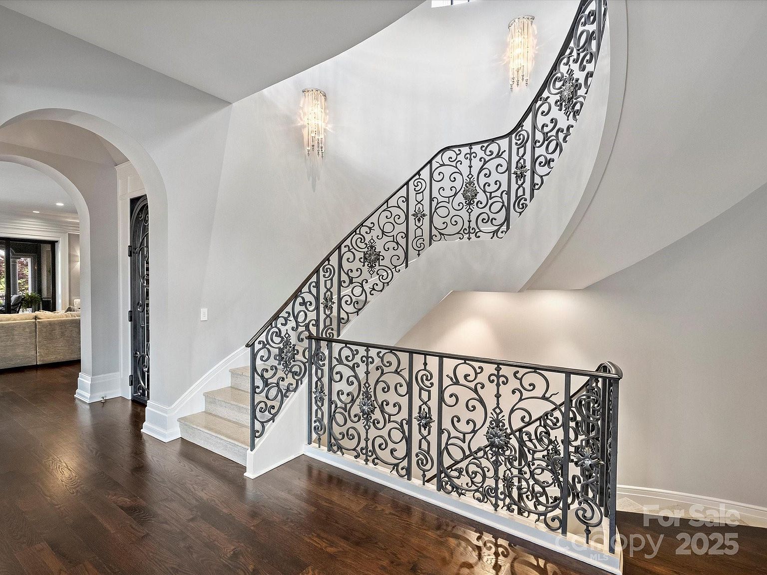This elegant entryway features a grand curved staircase with intricate wrought-iron railings and light-colored stone steps. The space is illuminated by two ornate crystal wall sconces mounted on a smooth, neutral-toned wall, while dark hardwood flooring provides a rich contrast. An arched doorway leads into an adjacent living area, creating an open and sophisticated architectural flow.