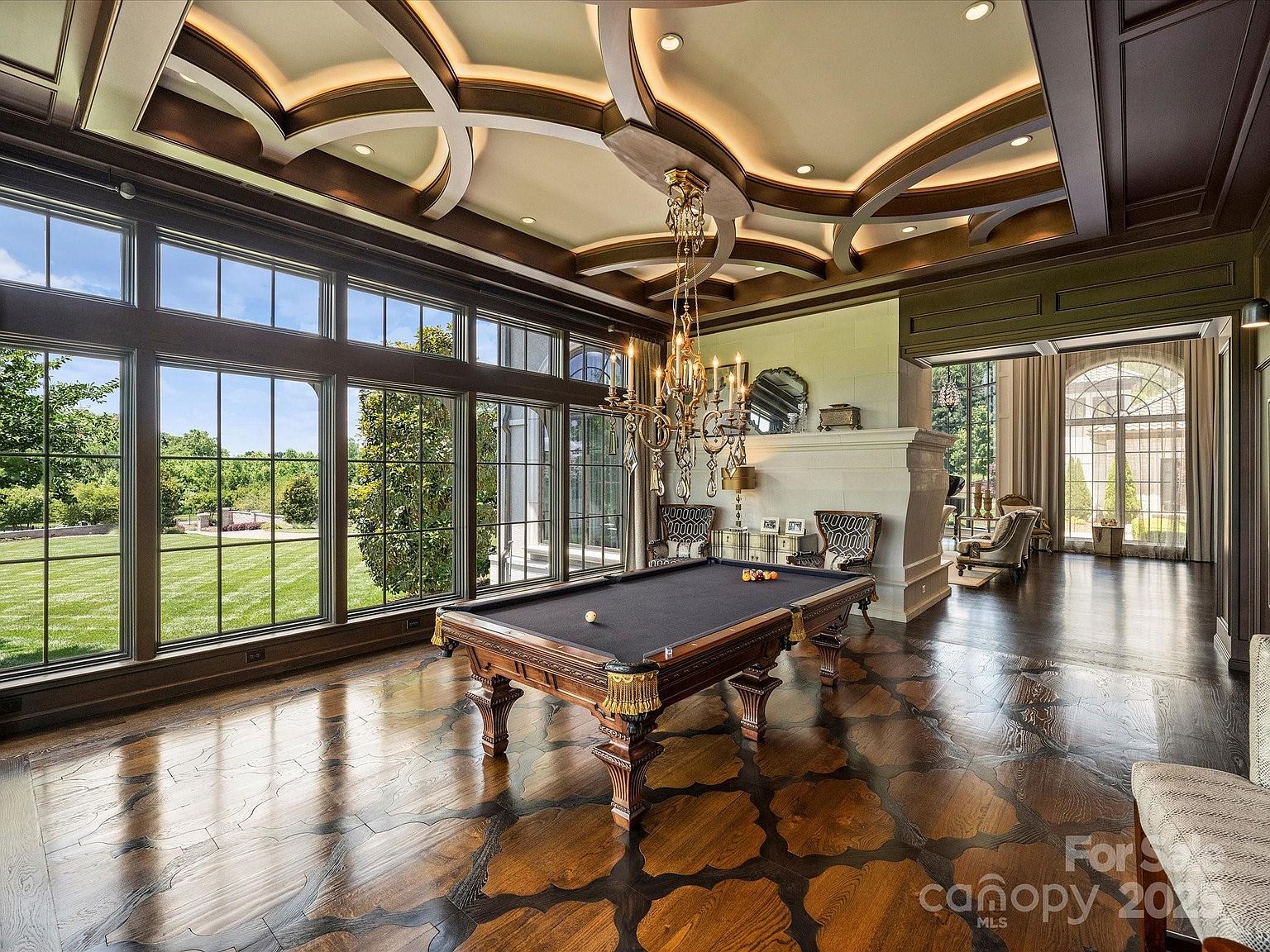 This elegant game room features a prominent billiards table as its centerpiece, set against a backdrop of expansive windows that offer a view of the lush green lawn outside. The room is defined by its intricate, dark-toned patterned wood flooring and a sophisticated coffered ceiling with recessed lighting. A tall, light-colored stone fireplace surround adds a sense of grandeur, while a classic crystal chandelier hangs above the table, creating a refined and luxurious atmosphere.