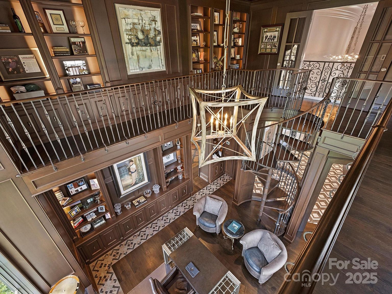 This high-angle, cinematic shot captures a sophisticated two-story library or study featuring rich, dark wood paneling and extensive built-in shelving. A striking geometric chandelier serves as the focal point above a central seating area with two leather armchairs and a desk, while a spiral staircase provides elegant access to the upper gallery. The space exudes a classic, scholarly atmosphere with curated decor and a patterned tile floor accentuating the lower level.
