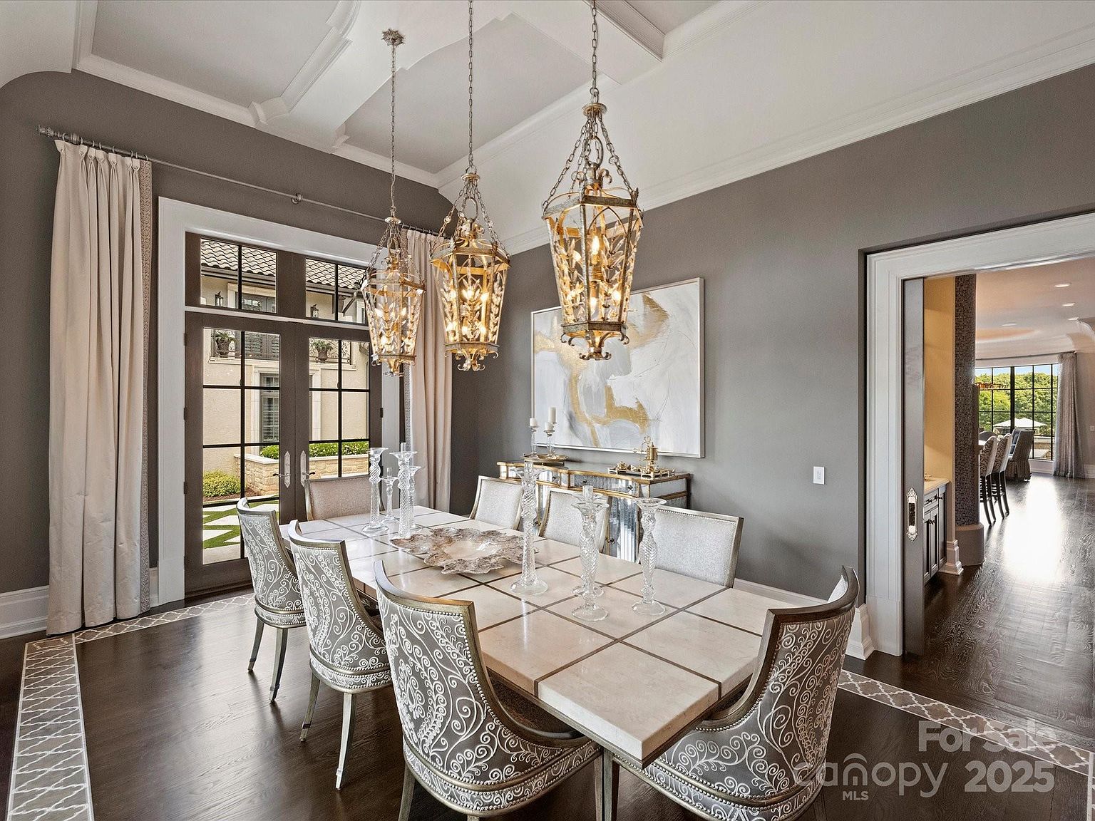 This elegant dining room features a large rectangular table with a stone-tiled top, surrounded by six chairs upholstered in a decorative, light-colored scroll pattern. Three ornate, multi-tiered metal lanterns hang from the ceiling, casting a warm glow over the space, while a mirrored sideboard sits against the gray wall beneath a large abstract painting. The room is framed by dark wood flooring, tall windows with light drapery, and a wide doorway leading to an adjacent hallway.