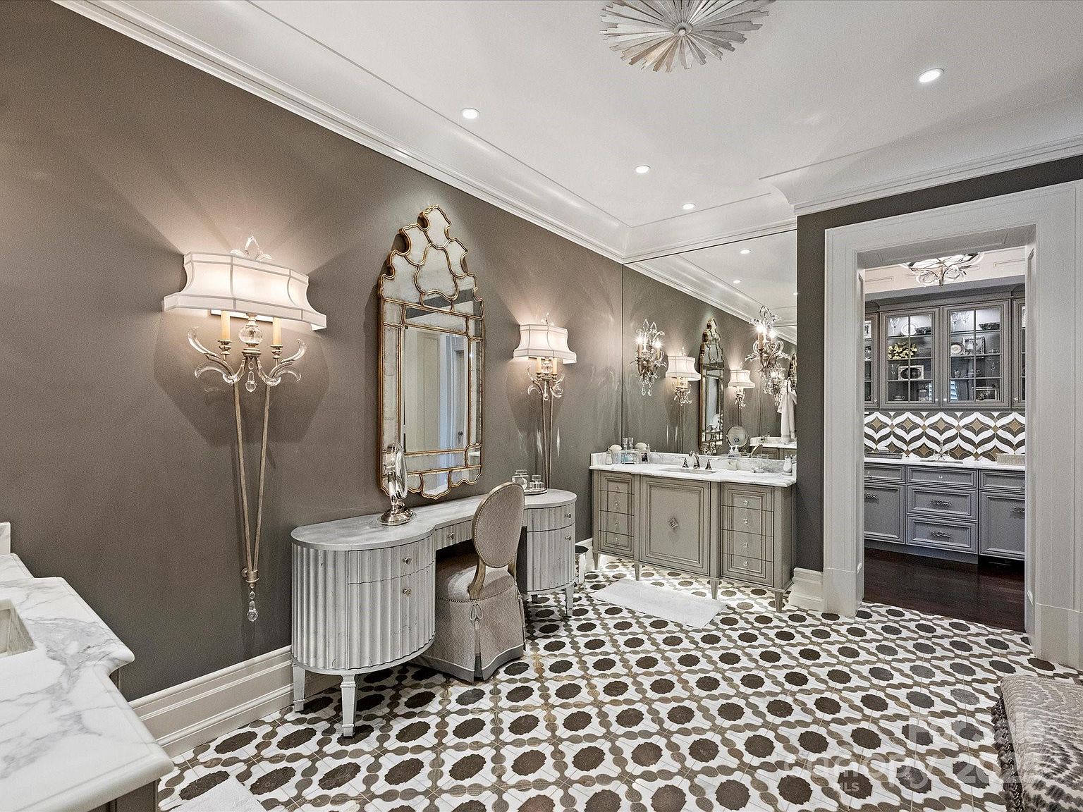 This elegant primary bathroom features a sophisticated vanity area with a decorative mirror, wall-mounted sconces, and a matching upholstered chair. The space is anchored by a patterned tile floor and leads into an adjacent room with additional cabinetry and a geometric backsplash. The overall aesthetic is refined and luxurious, characterized by neutral tones and high-end finishes.