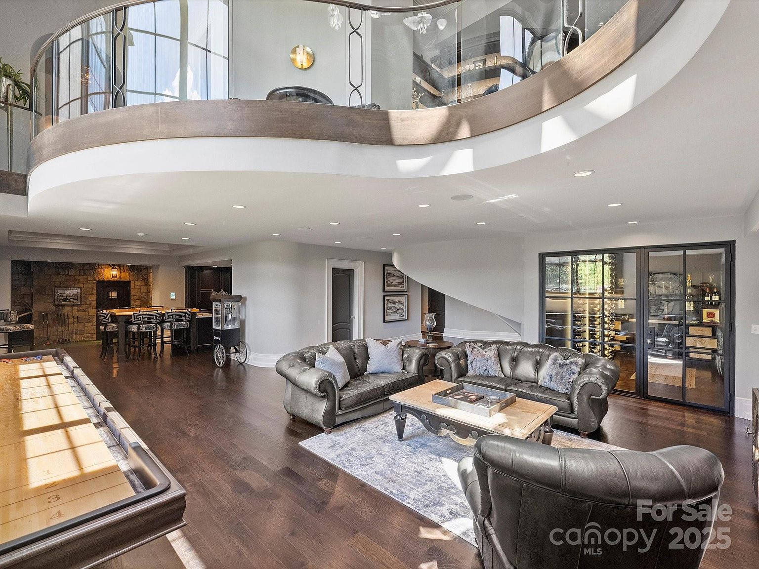 This expansive, open-concept basement features a sophisticated entertainment area with dark leather seating, a central coffee table, and a shuffleboard table in the foreground. To the left, a bar area with stone-clad walls and high-top seating provides a cozy lounge atmosphere, while a glass-enclosed wine cellar sits to the right. The space is defined by a dramatic curved mezzanine balcony above, creating an airy, multi-level architectural feel.