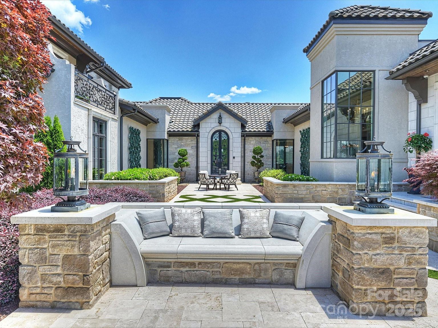 This elegant outdoor courtyard features a built-in stone bench with plush gray cushions, framed by two decorative lanterns on stone pillars. The space leads to a central patio area with a small bistro set, surrounded by manicured landscaping and stone retaining walls. The architecture showcases a sophisticated Mediterranean-inspired design with a tiled roof and arched entryway, creating a serene and luxurious atmosphere.