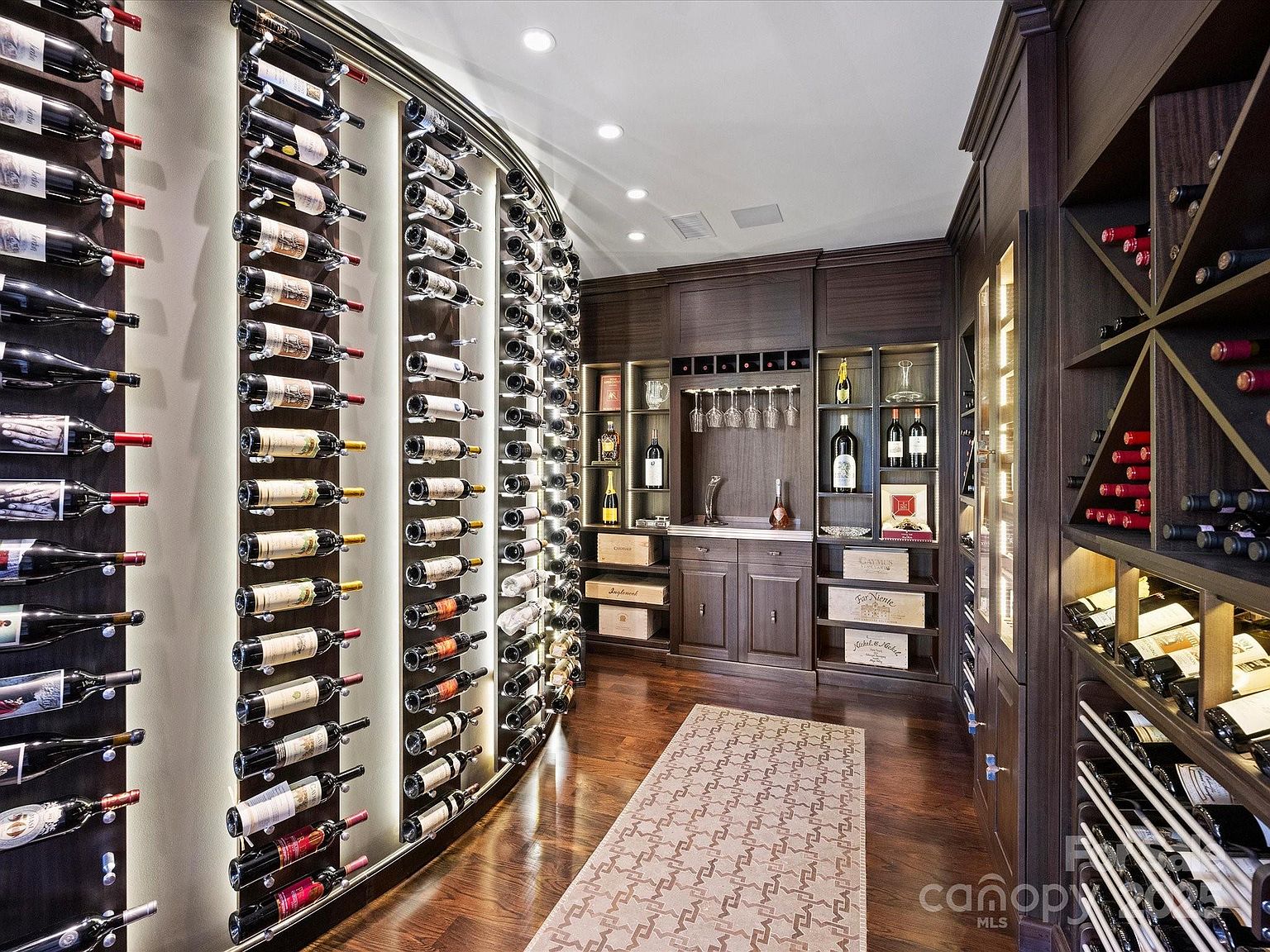 This sophisticated wine cellar features extensive custom dark wood cabinetry with integrated LED accent lighting that highlights the bottle displays. The room includes a curved wall of horizontal bottle racks, a central tasting area with a counter and stemware storage, and a patterned area rug centered on the hardwood floor. The perspective is a wide-angle shot looking down the length of the cellar, showcasing the organized storage and elegant atmosphere.