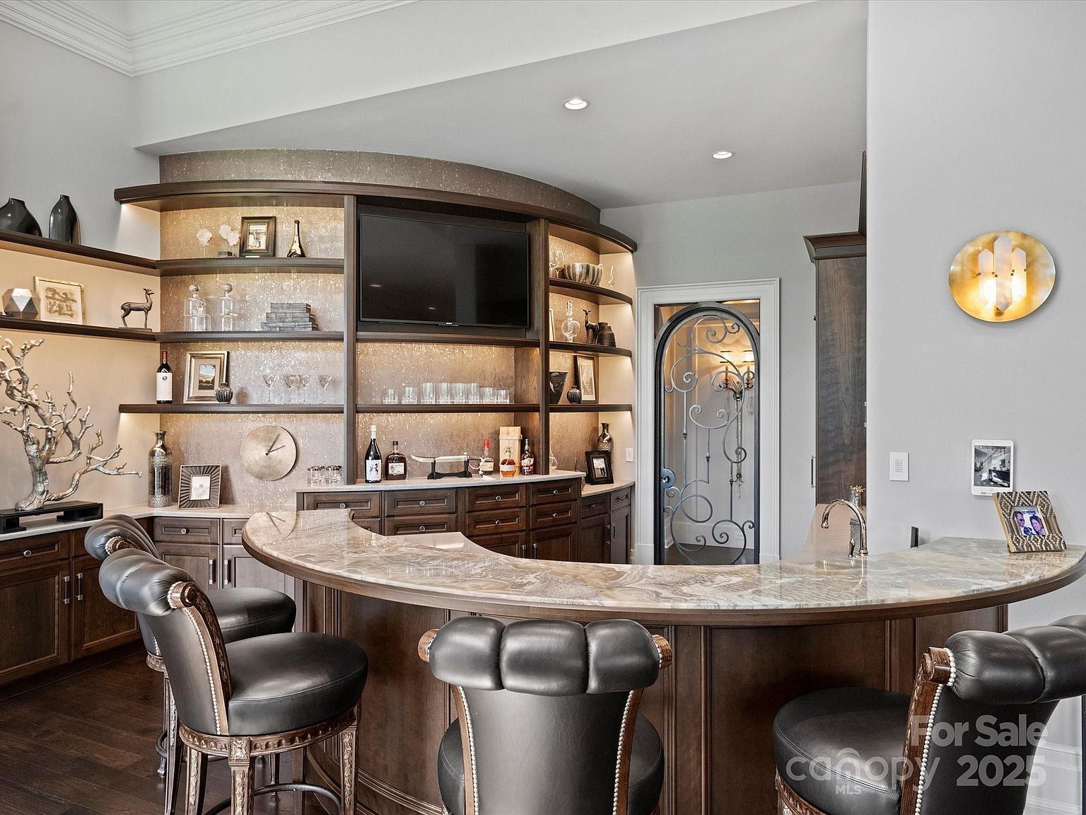 This sophisticated home bar area features a curved, stone-topped counter paired with dark leather bar stools. Behind the bar, a custom-built, curved shelving unit with integrated lighting displays glassware and decor, centered around a wall-mounted television. An arched, decorative metal door leads to an adjacent room, while a circular gold wall sconce adds a touch of elegance to the neutral-toned space.