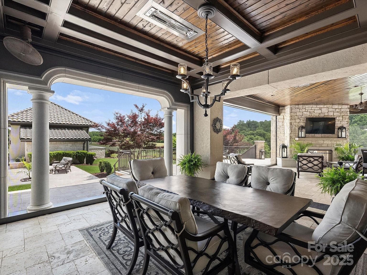 This expansive covered outdoor dining area features a rustic wood-paneled ceiling with exposed beams and a central chandelier. The space is furnished with a large dark wood table and cushioned chairs, set upon a stone-tiled floor that extends toward an open-air view of the landscaped grounds. A stone-clad chimney breast with an integrated television serves as a focal point in the background, creating a sophisticated and inviting atmosphere for al fresco entertaining.